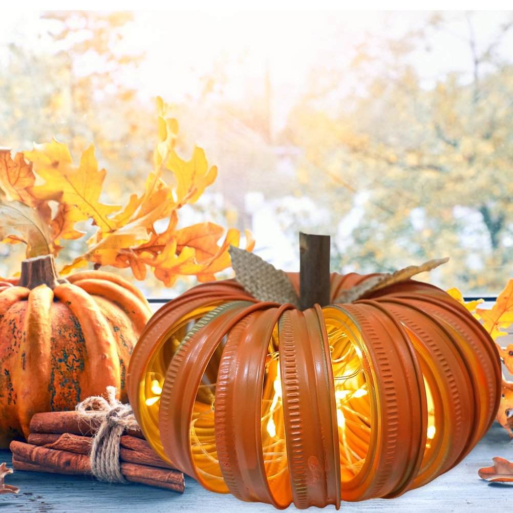 Ein orangefarbener Kürbis mit Lichterketten im Inneren, neben einem echten Kürbis und Zimtstangen. Herbstblätter im Hintergrund.