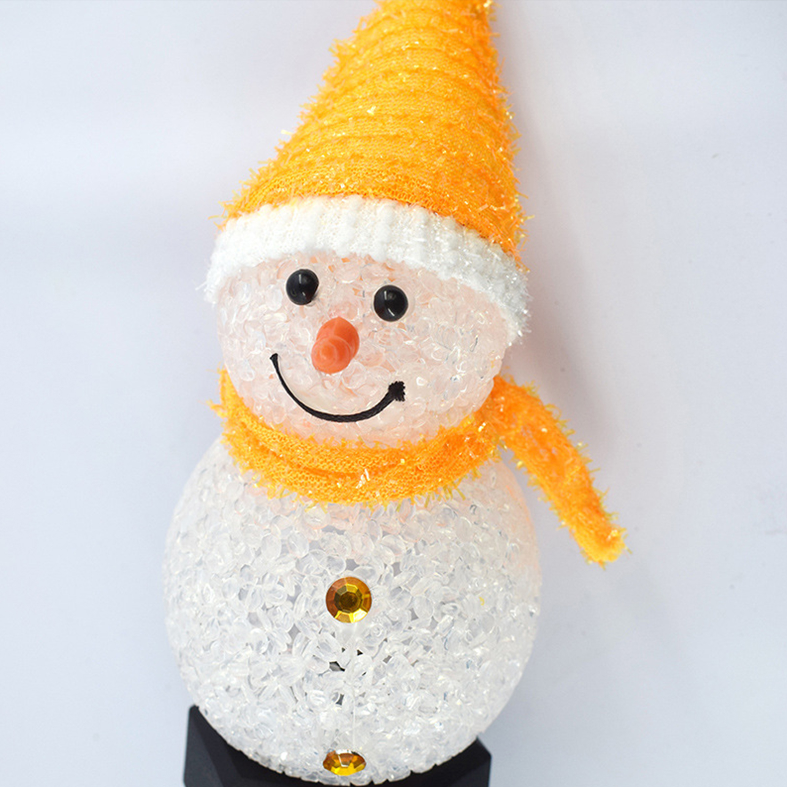 Eine Schneemann-Ornament mit gelber Mütze, Schal und Nase, schwarzen Augen und Lächeln, und einem Zierstein am Körper.