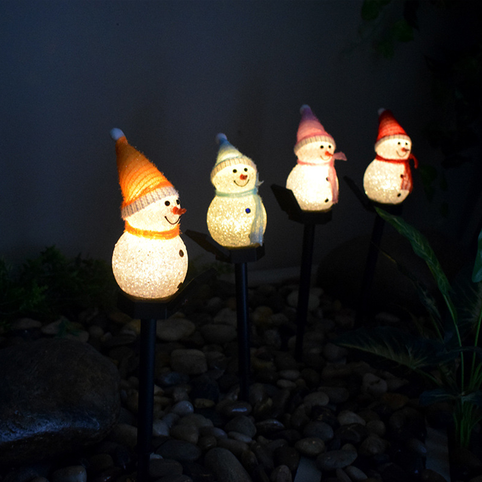 Vier leuchtende Schneemann-Gartenlichter. Jeder trägt einen Hut und Schal in einer anderen Farbe. Auf einem Steinhaufen.