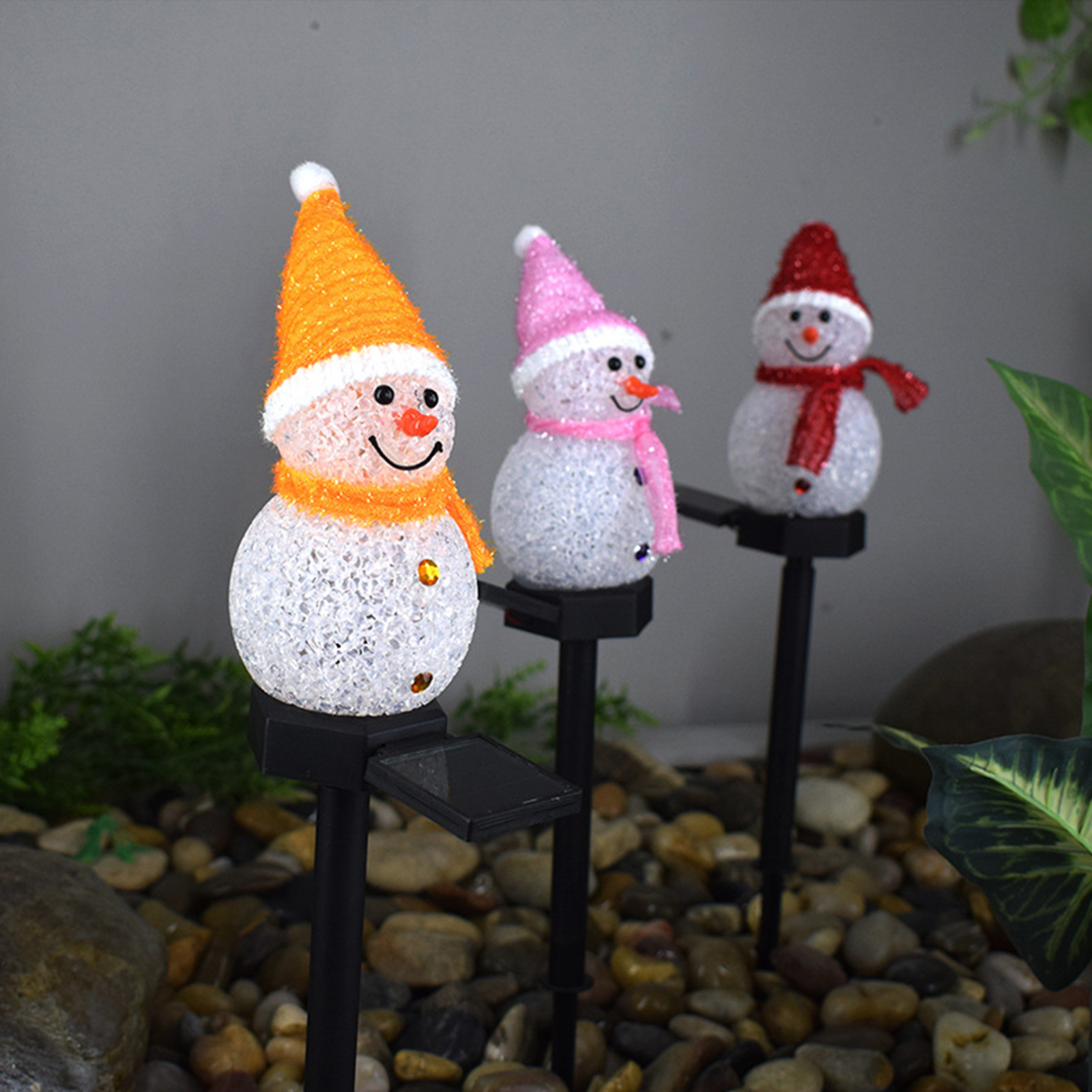 Drei Schneemann-Dekorationen mit orangen, rosa und roten Hüten und Schals. Sie stehen auf schwarzen Pfählen in einem Beet.