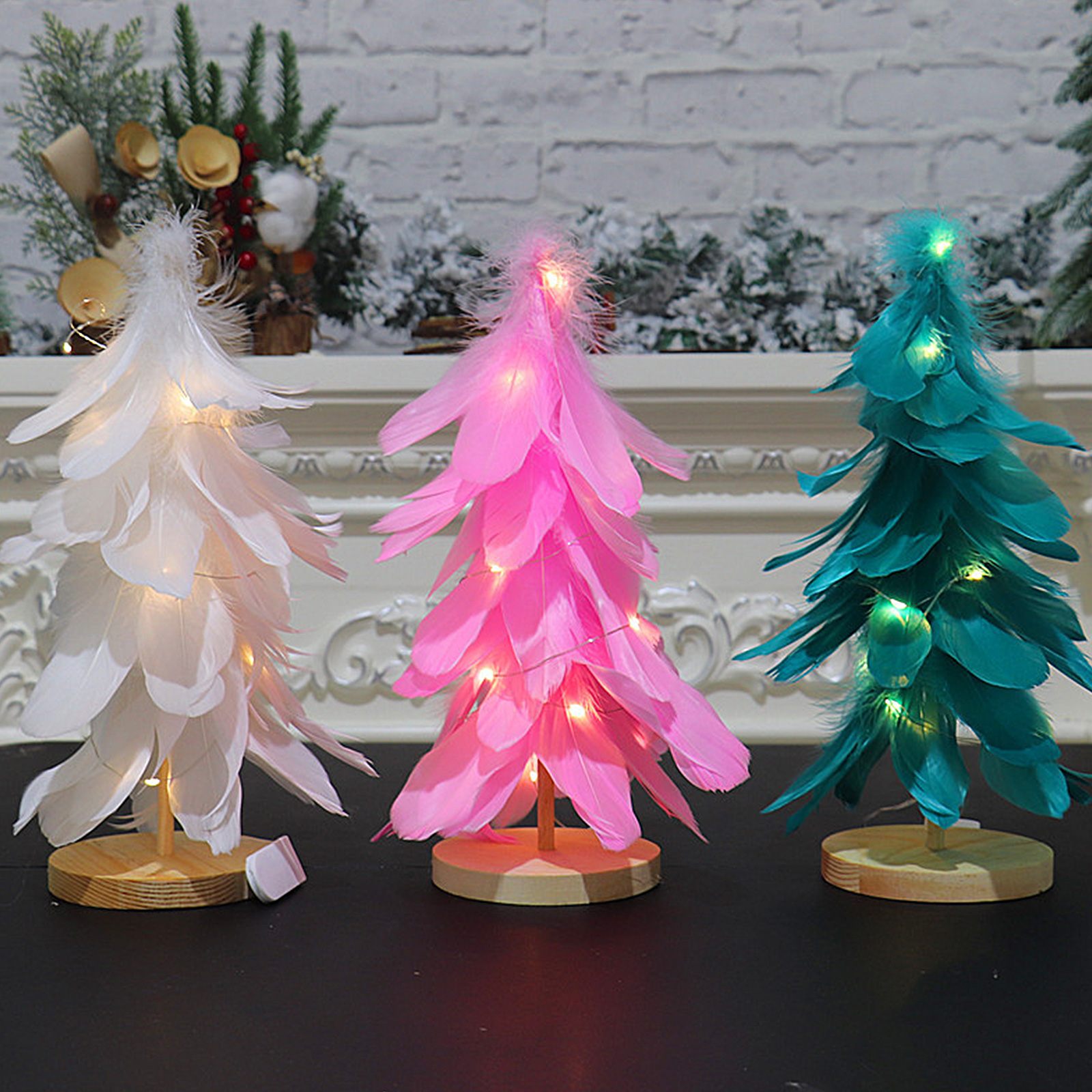 Drei Weihnachtsbaumfedern in Weiß, Rosa und Türkis, jede auf Holzsockel, mit Feenlichtern beleuchtet, auf einer schwarzen Oberfläche.