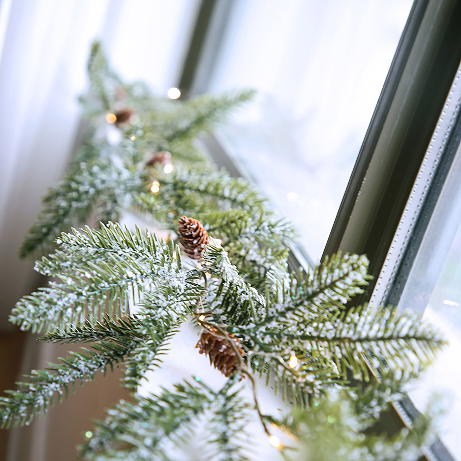 Weihnachts-Girlande am Fenster. Grüne Tannenzweige mit weißem Schnee und braunen Tannenzapfen. Kleine Lichter sind an.