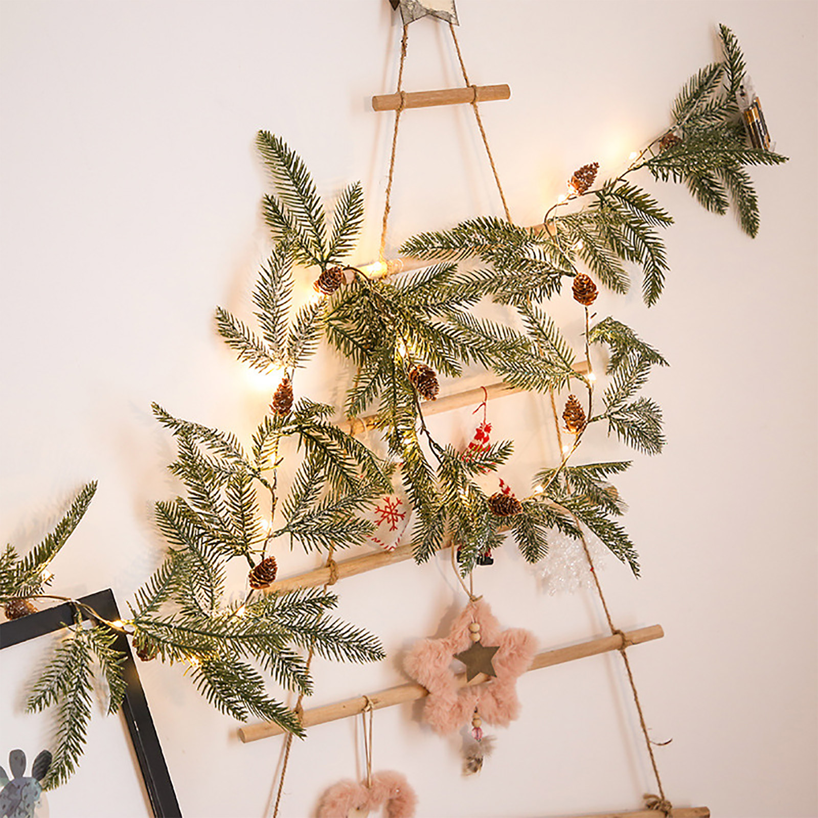 Eine leiterförmige Weihnachtsdekoration mit Tannenzweigen, Tannenzapfen, Lichtern und Ornamenten hängt an einer weißen Wand.