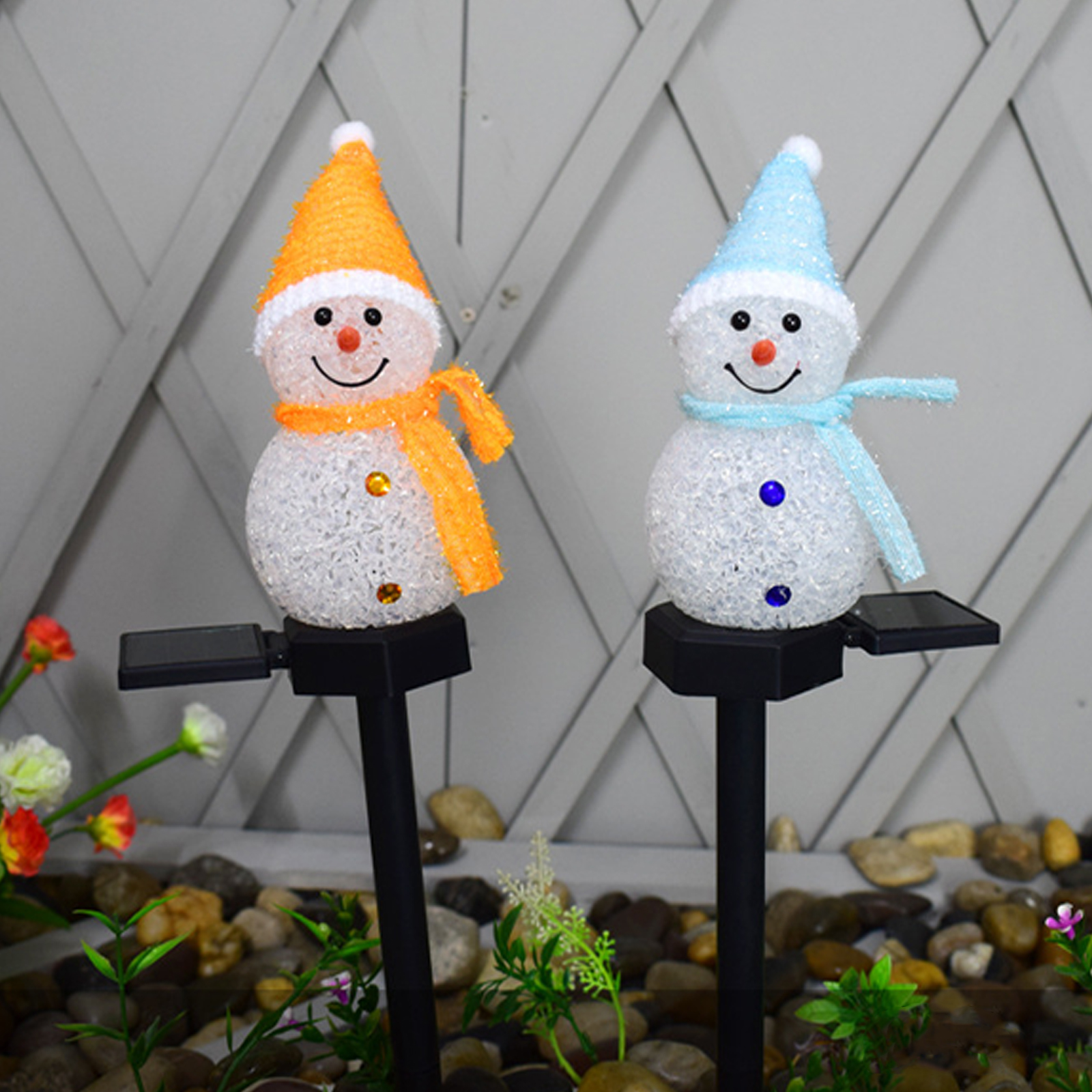 Zwei Schneemann-Gartenlichter, eines mit orangen, das andere mit blauen Akzenten. Sie haben Hüte, Schals und Lächeln. Steine und Pflanzen sind im Hintergrund.