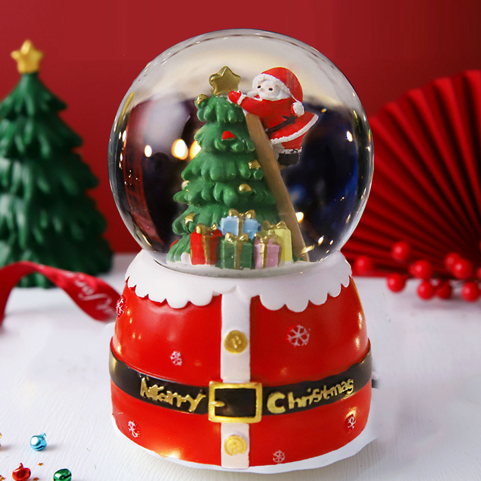 Eine Schneekugel mit einem Weihnachtsmann, der einen Weihnachtsbaum schmückt. Sie steht auf einem roten Santa-Anzug-Sockel. Andere Weihnachtsdekorationen im Hintergrund.