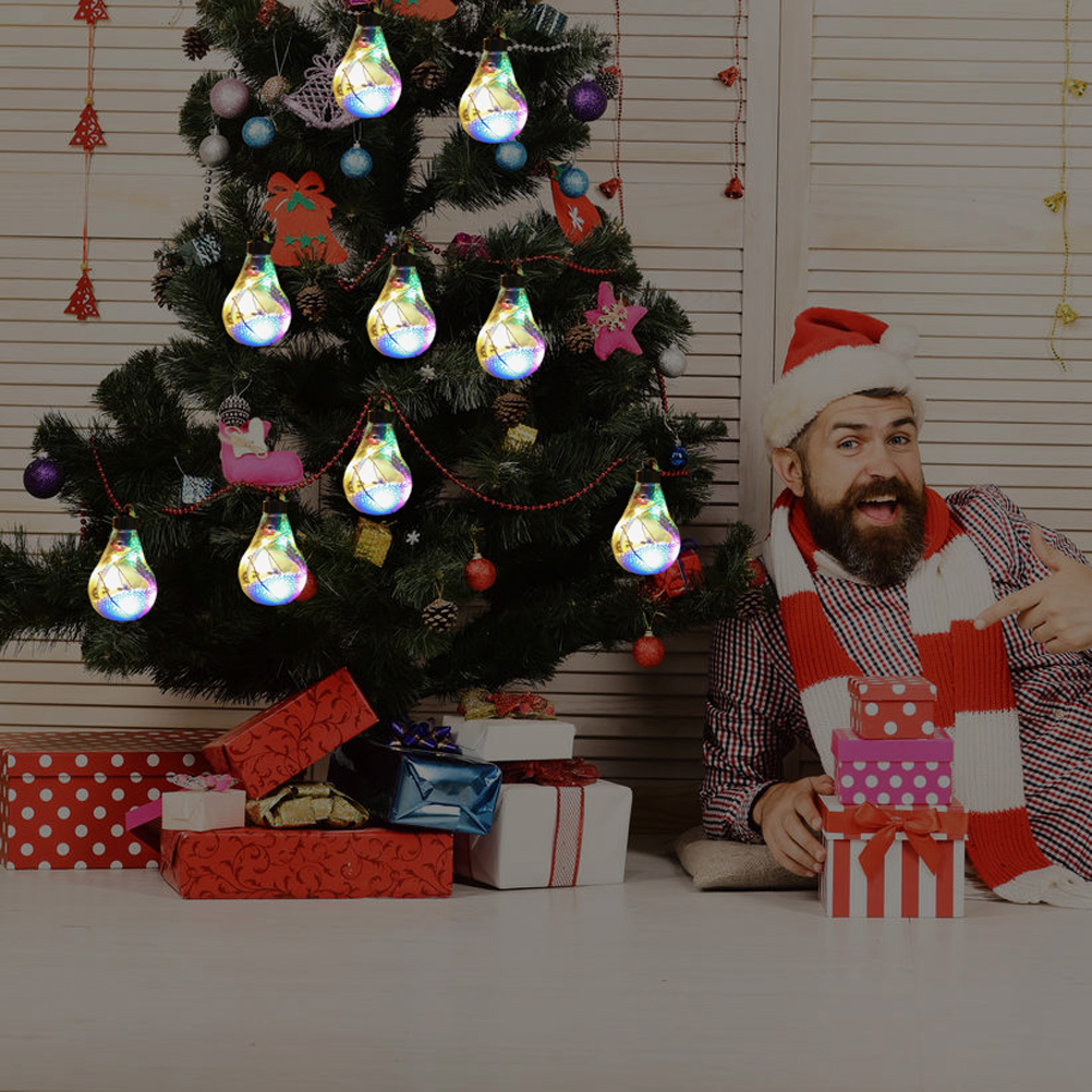 Ein Mann mit Weihnachtsmütze und Schal zeigt auf einen Weihnachtsbaum mit Glühbirnen. Geschenke liegen unter dem Baum.