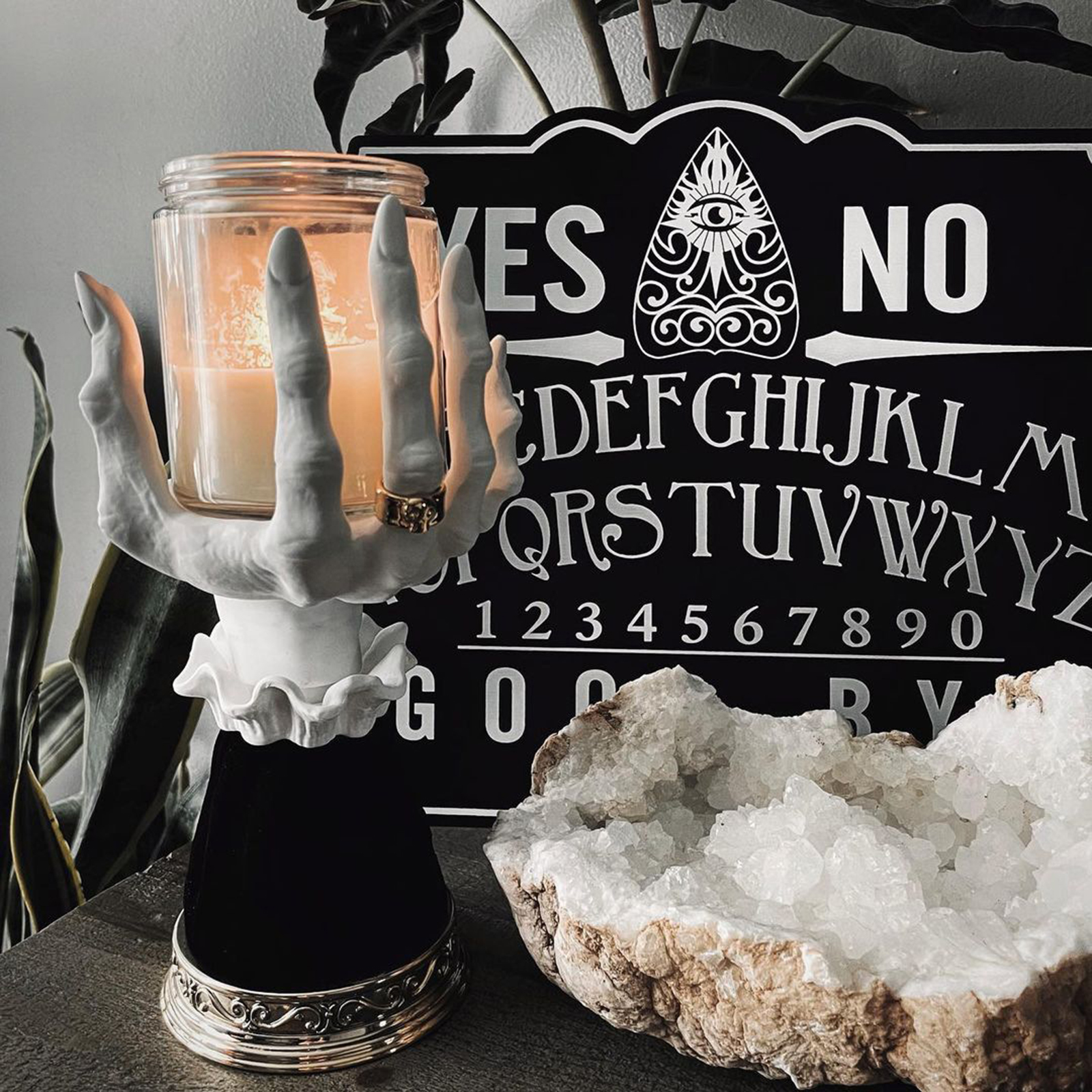Eine Kerze in einem Glas wird von einer weißen Handstatue gehalten, mit einem Ouija-Brett im Hintergrund und einer Geode auf dem Tisch.