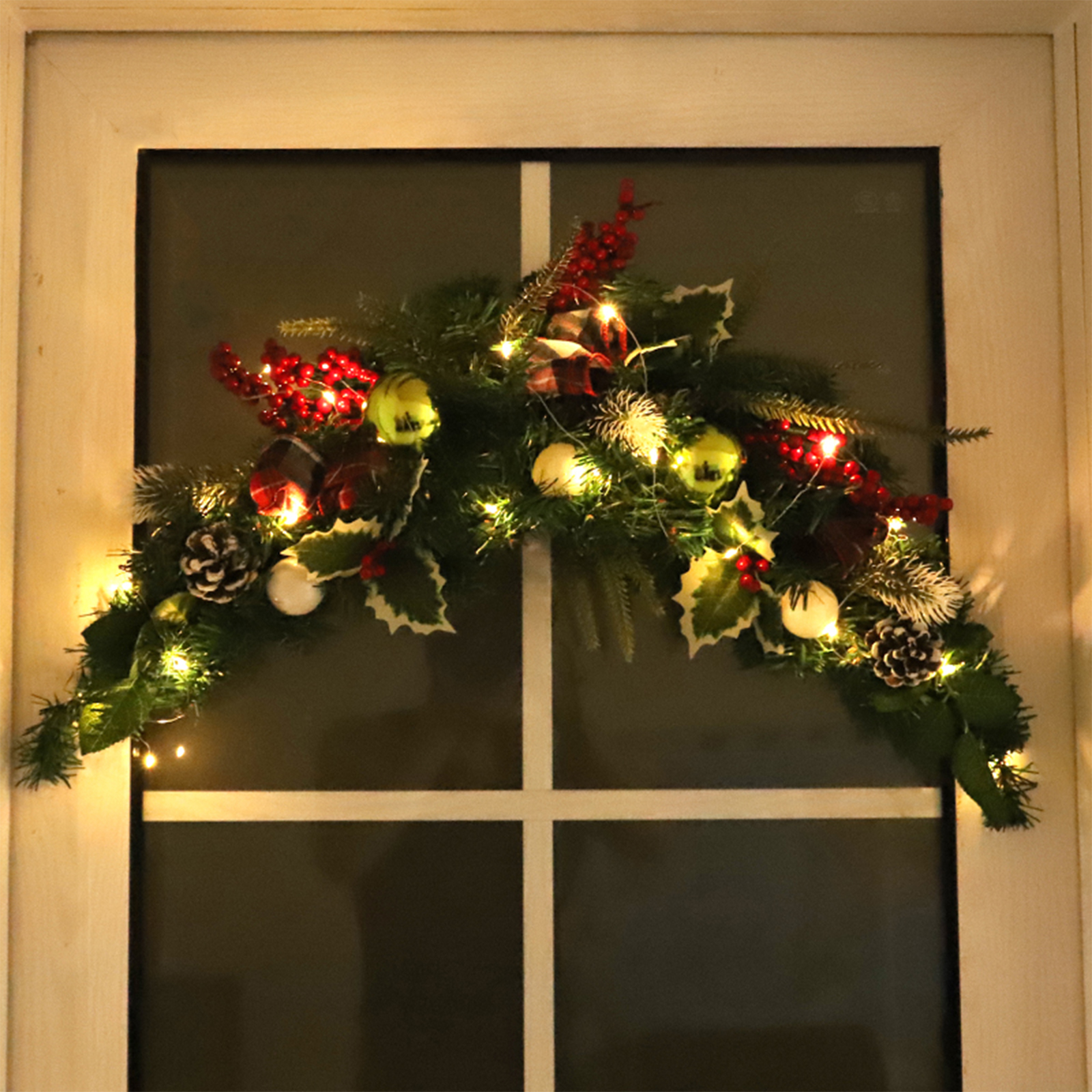 Ein Fenster mit einem Weihnachtsgirlande geschmückt. Es hat Lichter, Beeren, Ornamente und Tannenzapfen.