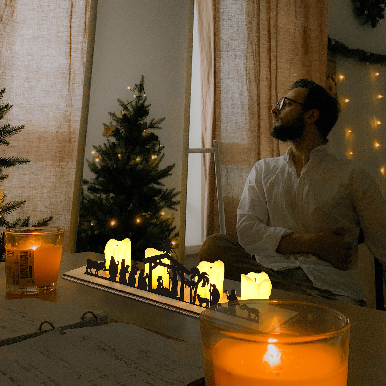 Ein Mann blickt nach oben in der Nähe einer von Kerzen beleuchteten Krippenszene. Weihnachtsbaum und Kerzen im Vordergrund. Warme, weiche Beleuchtung.