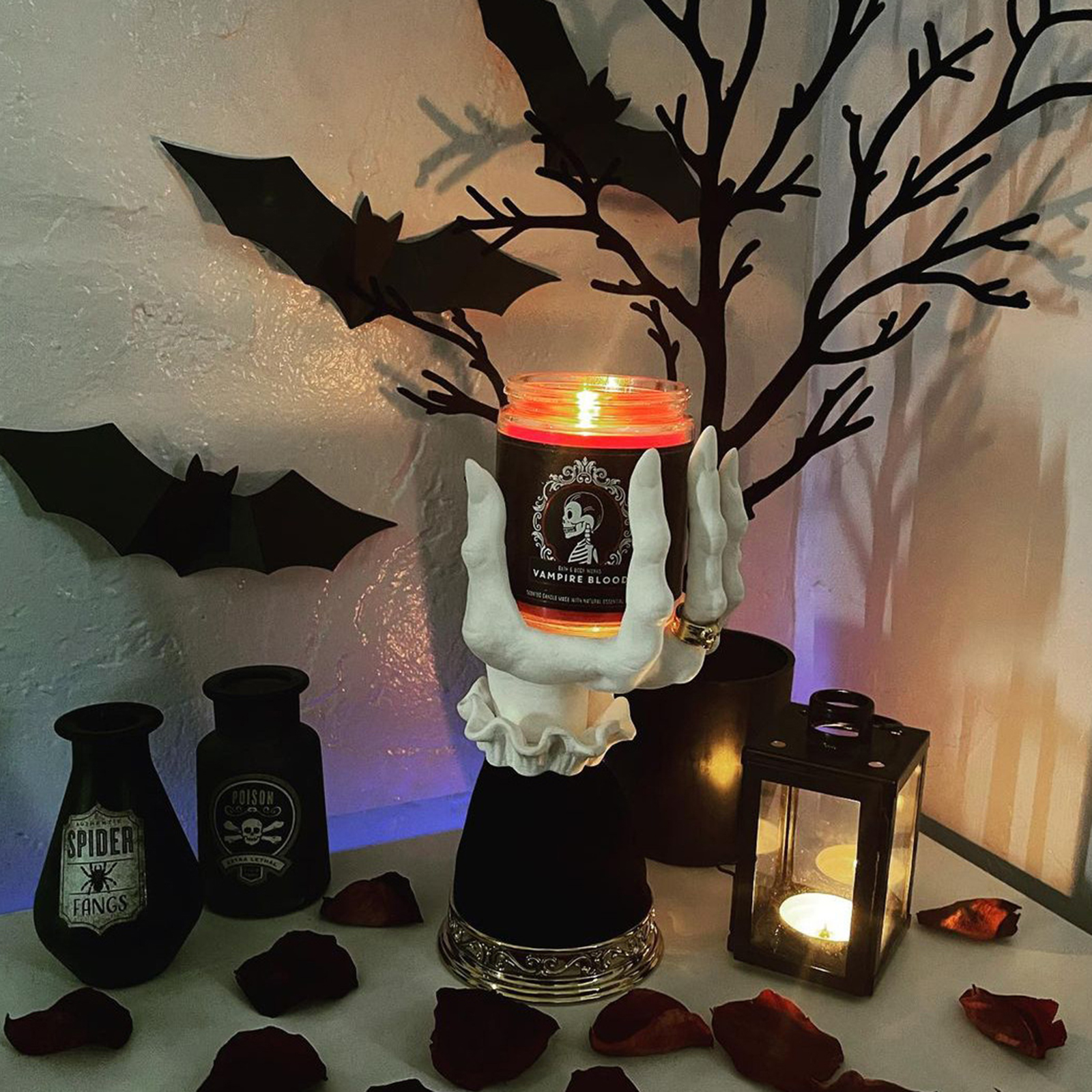 Halloween-Szene mit Fledermäusen, einer Kerze in einer Hand, Gläsern und einer Laterne. Rote Rosenblätter sind verstreut.
