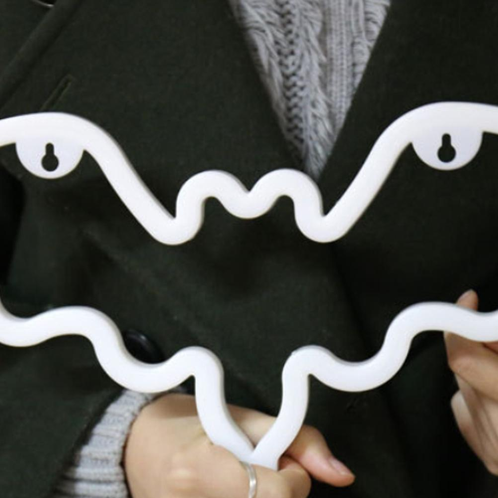 Eine Person hält einen weißen, wellenförmigen Kleiderbügel. Der Bügel hat ein gesichtsähnliches Design mit zwei Augenlöchern und einer Mundform. Der Hintergrund ist ein dunkler Mantel.