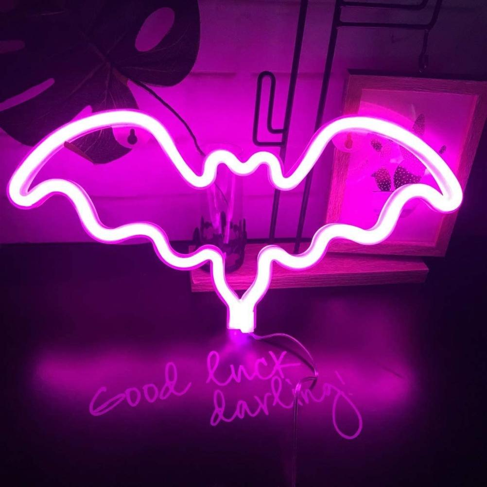 Ein neonfarbenes Fledermausschild leuchtet rosa mit dem Schriftzug 'Good luck darling'. Im Hintergrund sind ein Bild und eine Pflanze zu sehen.