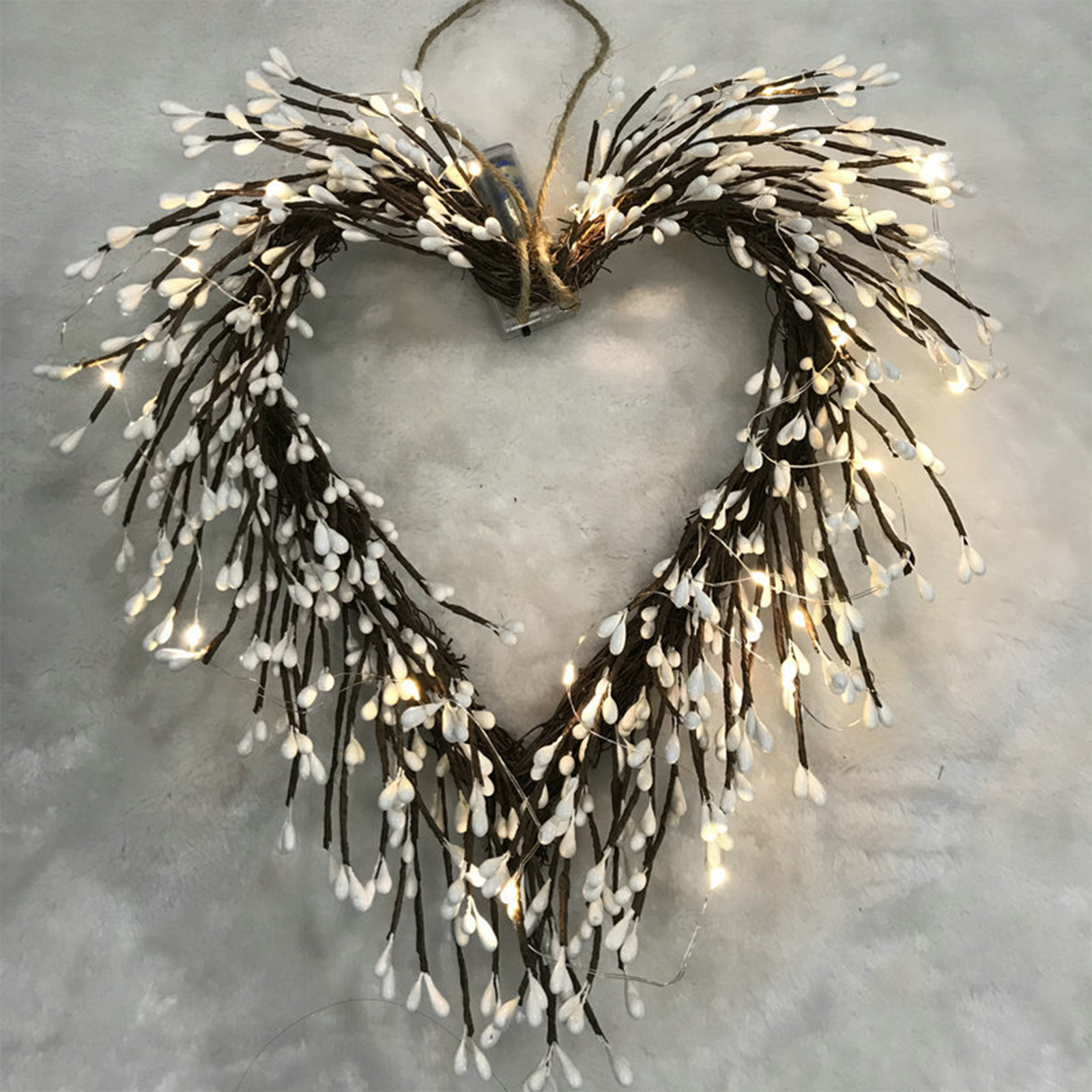Ein herzförmiger Kranz aus braunen Zweigen und weißen, tropfenförmigen Ornamenten, beleuchtet von kleinen Lichtern, hängt an einer hellgrauen Oberfläche.