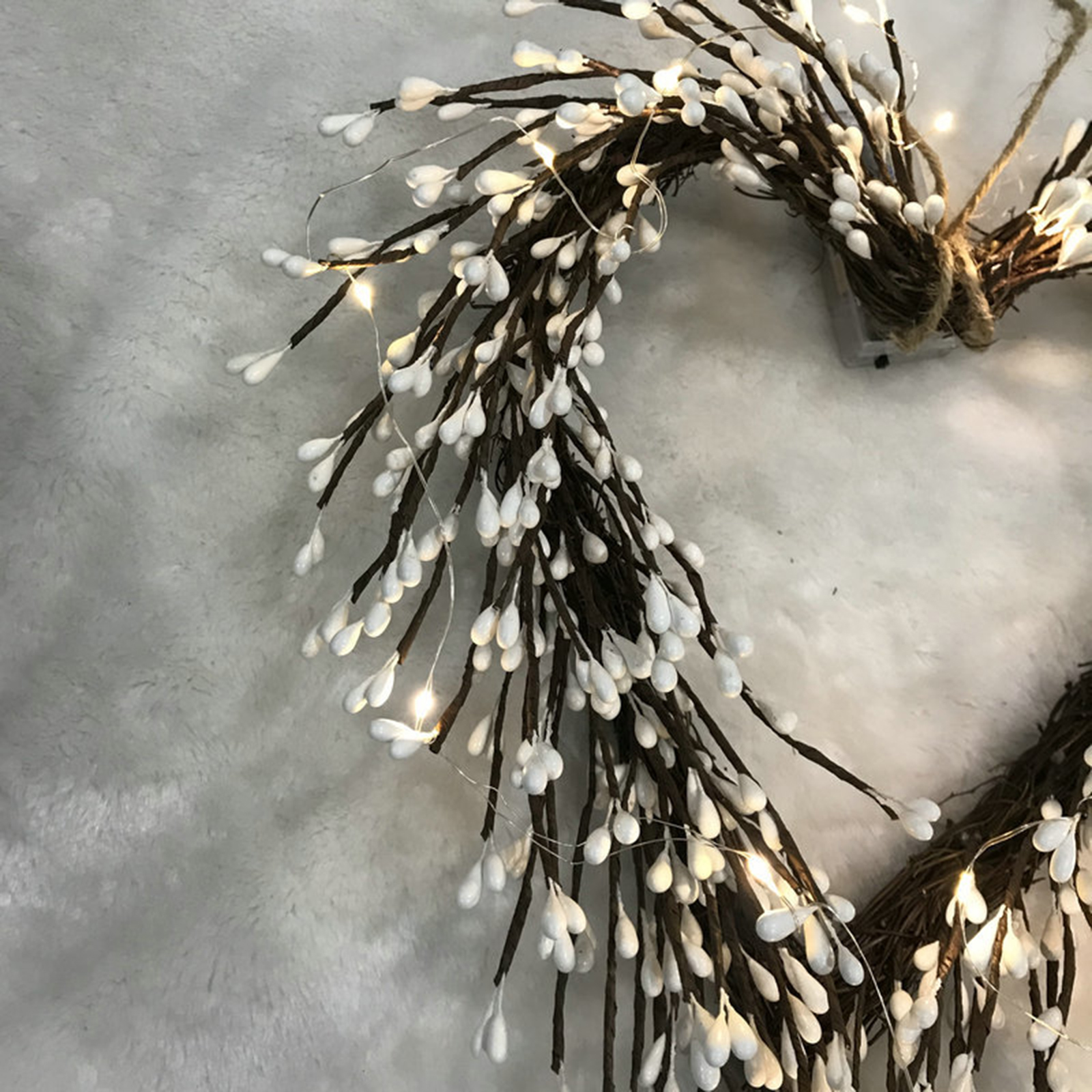 Ein herzförmiger Kranz aus braunen Zweigen mit weißen, tropfenförmigen Ornamenten, beleuchtet von kleinen Lichterketten auf weißem, flauschigem Hintergrund.