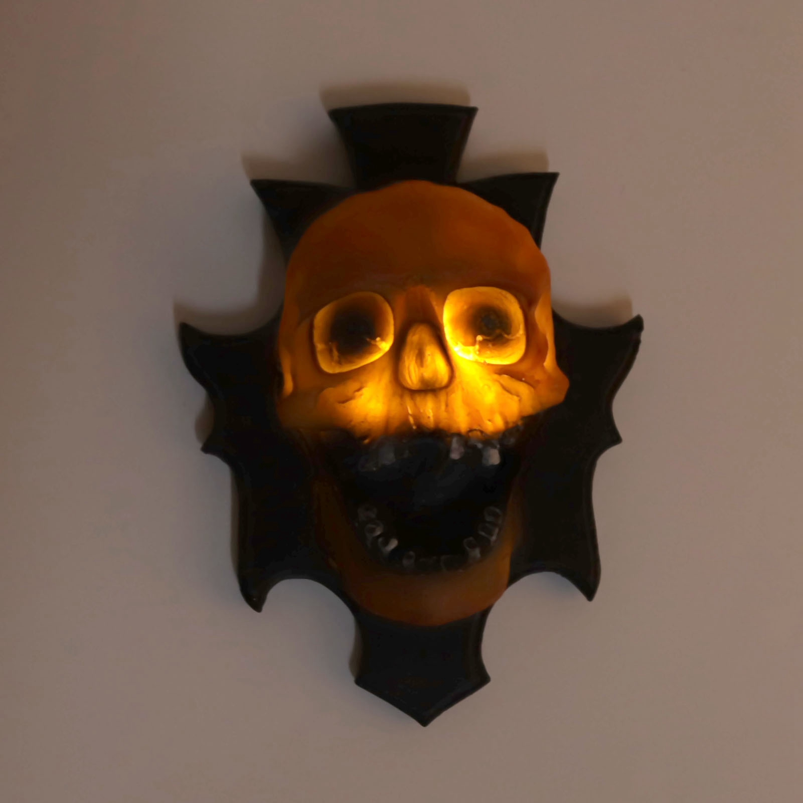Eine beleuchtete Totenkopfdekoration mit kreuzartigem Design, an einer Wand befestigt. Der Schädel ist orange mit leuchtenden Augen. Der Hintergrund ist weiß.
