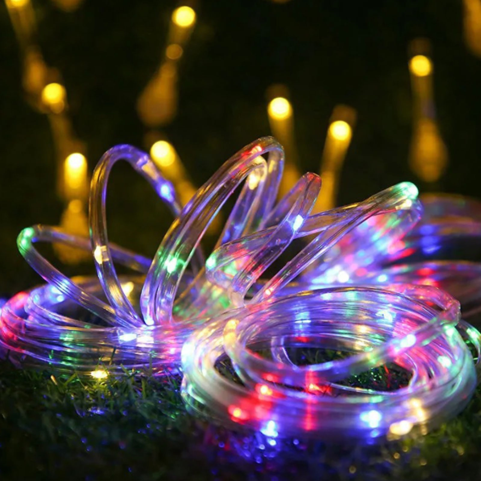 Nahaufnahme von bunten LED-Lichterketten, auf grünem Gras gerollt und ausgebreitet, mit verschwommenen gelben Lichtern im Hintergrund.