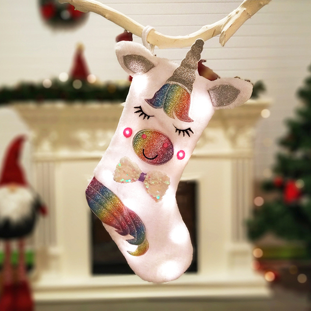 Ein Weihnachtsstrumpf in Form eines Einhorns, der an einem Ast hängt. Im Hintergrund befinden sich ein Kamin, ein Weihnachtsbaum und Dekorationen.