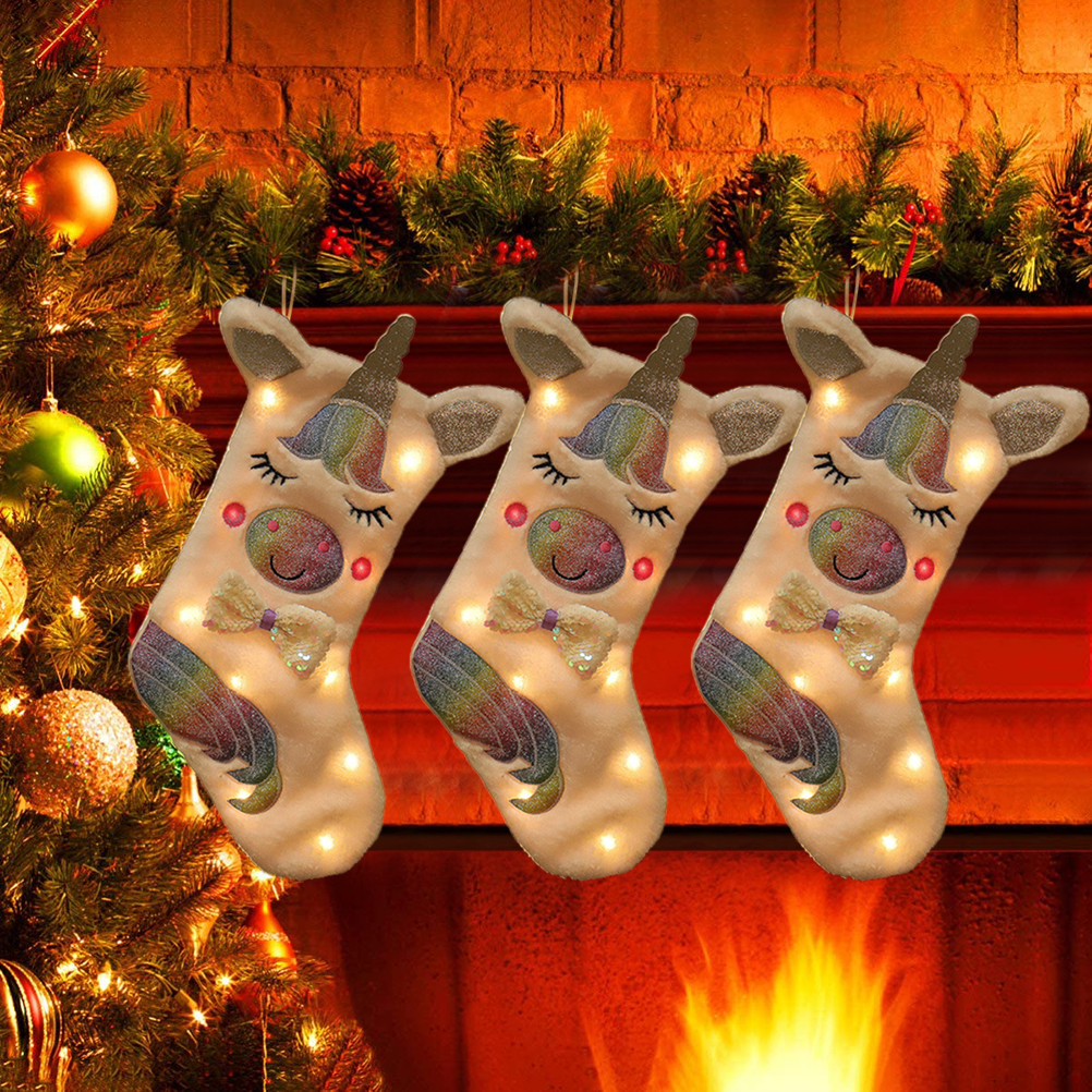 Drei Einhorn-Weihnachtsstrümpfe mit Lichtern hängen über einem Kamin. Links ist ein Weihnachtsbaum.
