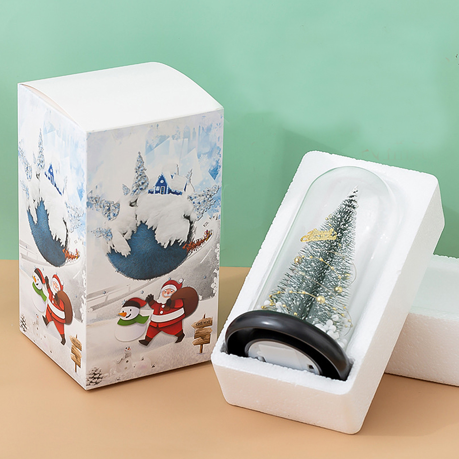 Ein Weihnachtsbaum in einer Glaskuppel neben einer Schachtel mit Santa- und Schneemann-Illustrationen.