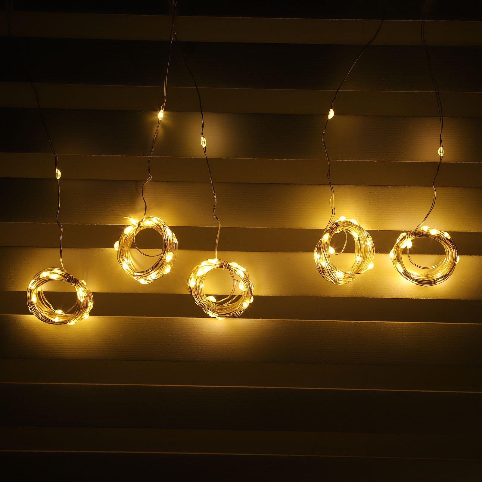 Fünf hängende, beleuchtete, kreisförmige Lichtdekorationen. Sie hängen vor einem hölzernen, lamellenförmigen Hintergrund. Die Lichter sind warmweiß.