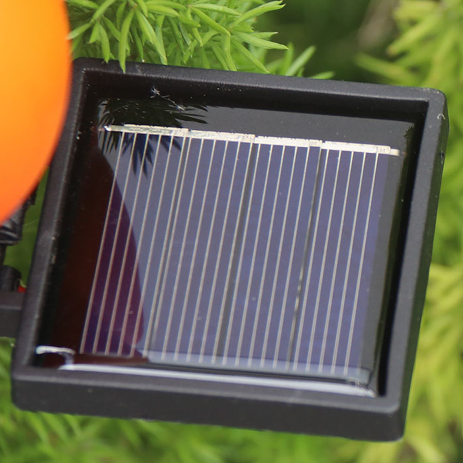 Nahaufnahme eines Solarpanels in einem schwarzen Rahmen, vor grünem Laub und einer orangefarbenen Kugel.