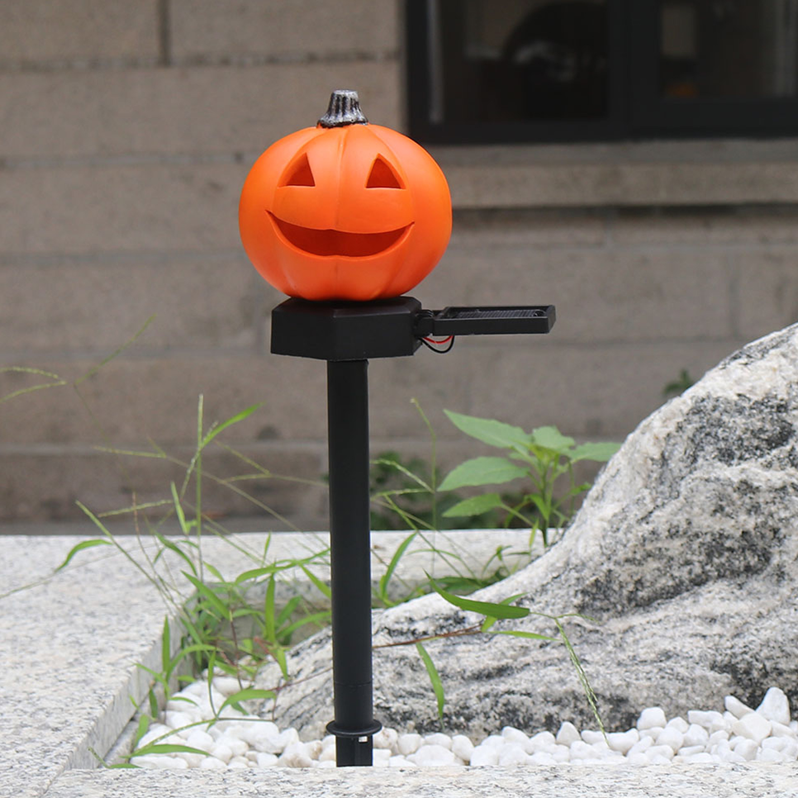 Eine orangefarbene Halloween-Kürbisdekoration auf einem schwarzen Pfahl, der im Freien mit Stein und Pflanzen aufgestellt ist.