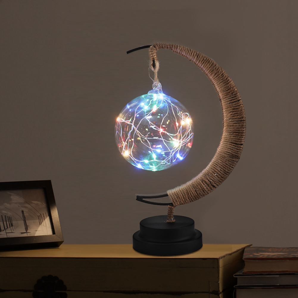 Eine dekorative Lampe mit einer leuchtenden Kugel aus Lichtern. Sie hat eine halbmondförmige Halterung und steht auf einem Tisch.