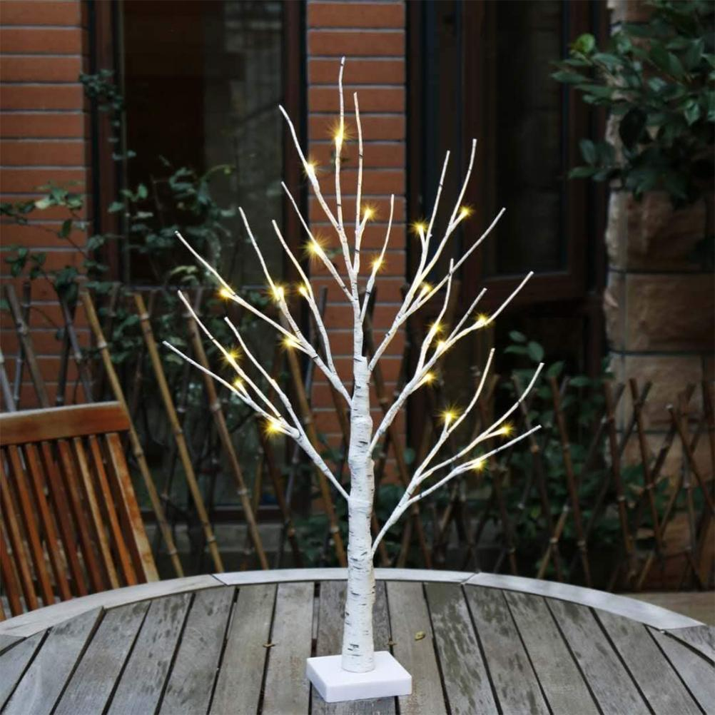 Ein kleiner weißer dekorativer Baum mit gelben Lichtern steht auf einem Holztisch. Im Hintergrund eine Ziegelmauer und Grün.