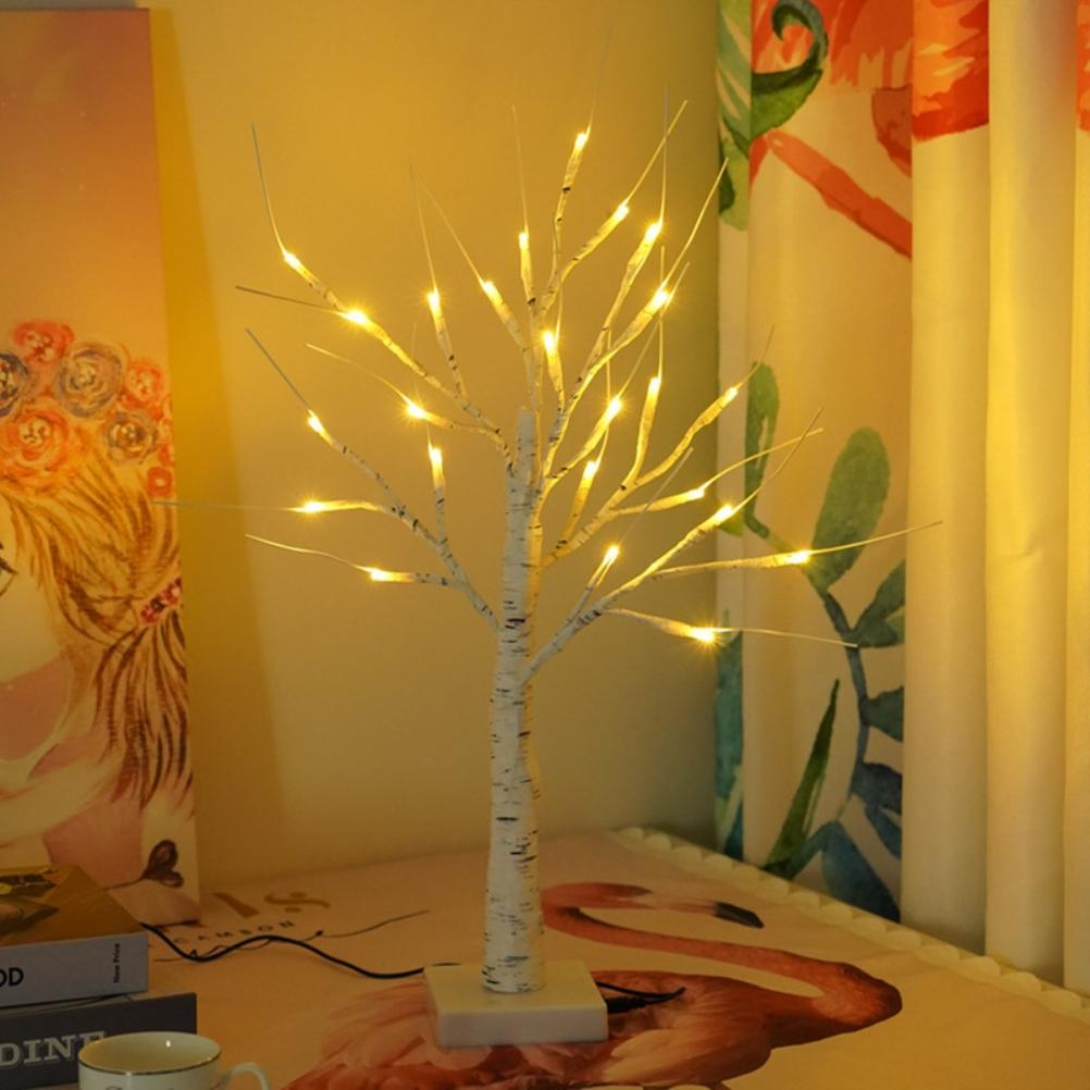 Eine kleine, beleuchtete Birkenbaumlampe auf einem Tisch mit floralem Muster und Vorhängen im Hintergrund. Bücher und eine Tasse sind sichtbar.