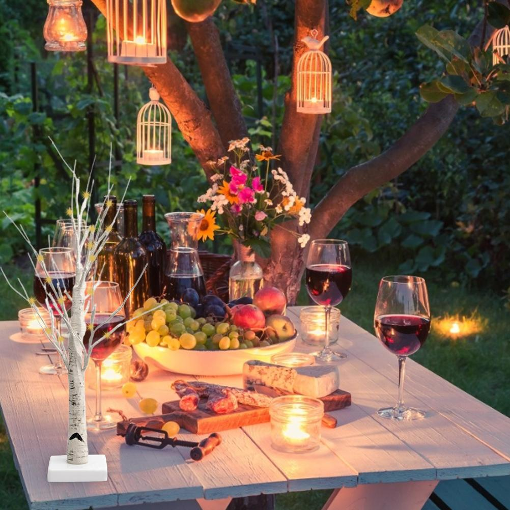 Ein Tisch ist für einen romantischen Abend gedeckt, mit Weingläsern, Obst, Käse und brennenden Kerzen unter einem Baum mit Laternen.