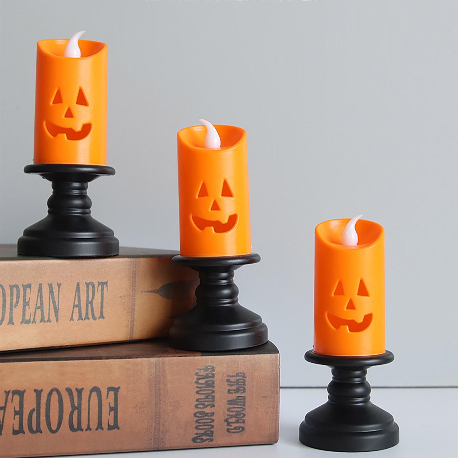 Drei orangefarbene Halloween-Kerzen mit geschnitztem Kürbisgesicht stehen auf schwarzen Ständern. Sie stehen auf Büchern.