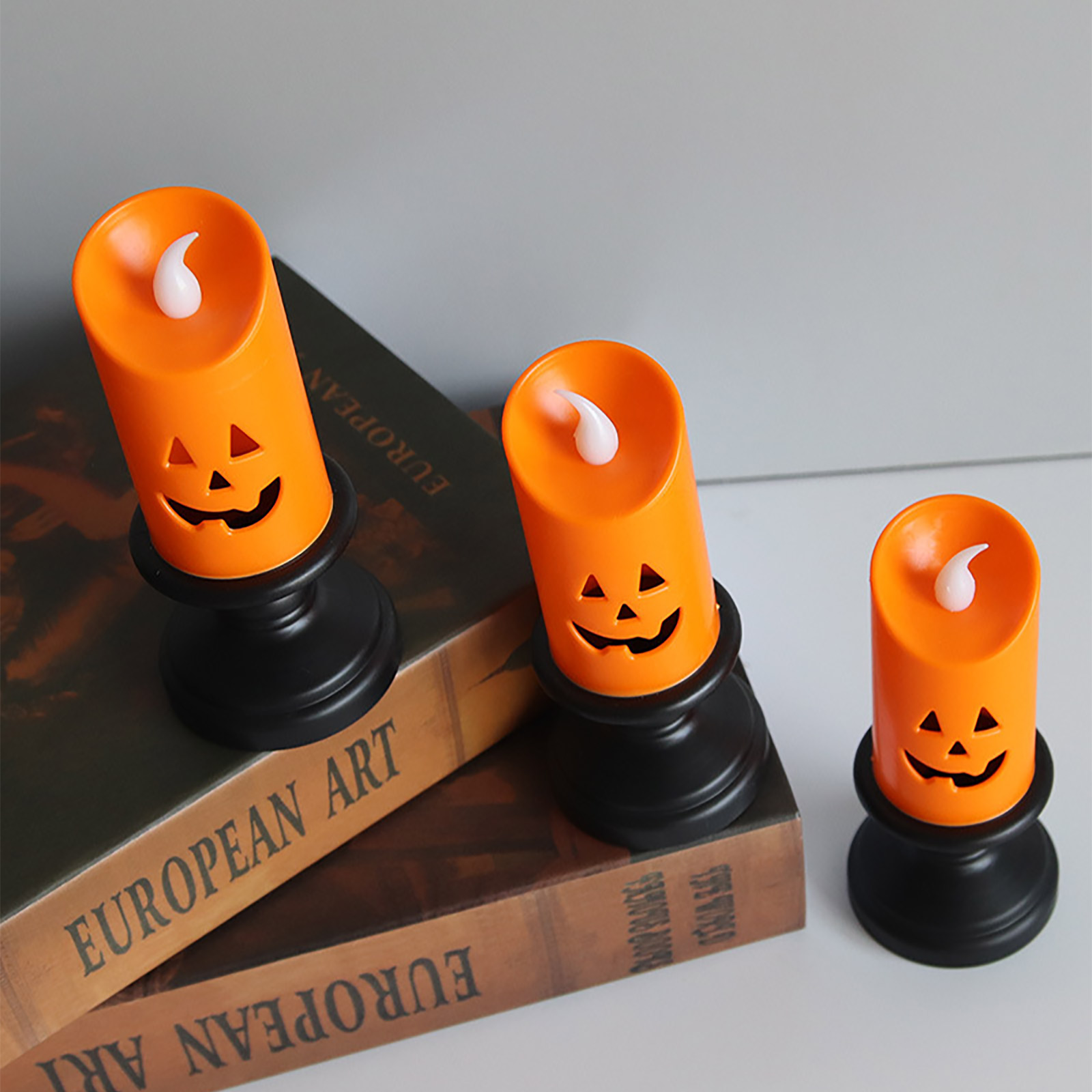 Drei orangefarbene Halloween-Kerzendekorationen mit Kürbisgesichtern auf schwarzen Sockeln. Sie stehen auf einem Buch mit der Aufschrift 'European Art'.