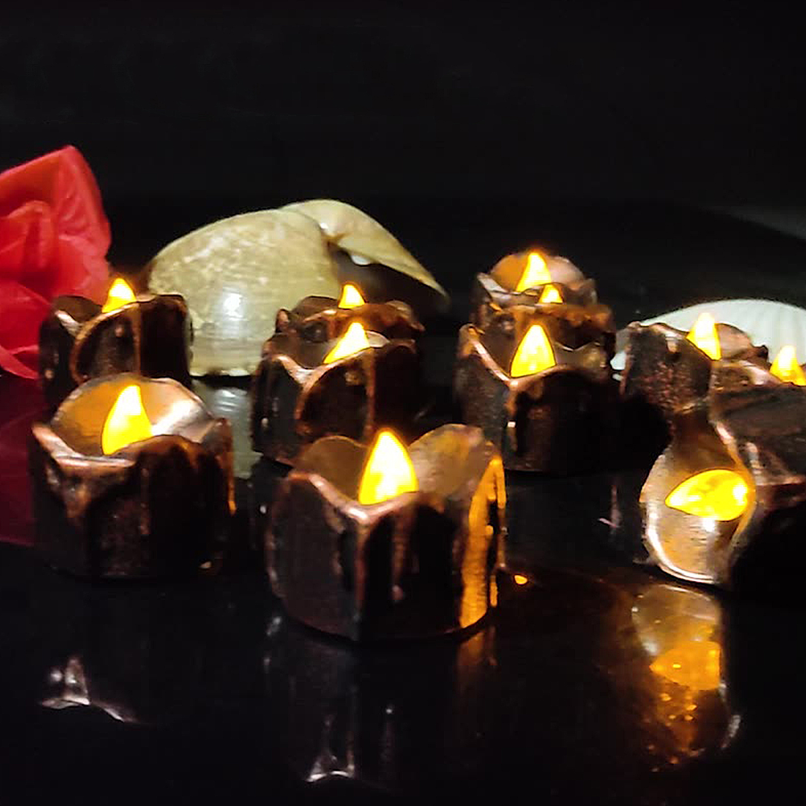 Mehrere braune, kerzenförmige Lichter mit gelb leuchtenden Flammen. Eine Muschel und roter Stoff sind im Hintergrund auf dunkler Oberfläche.