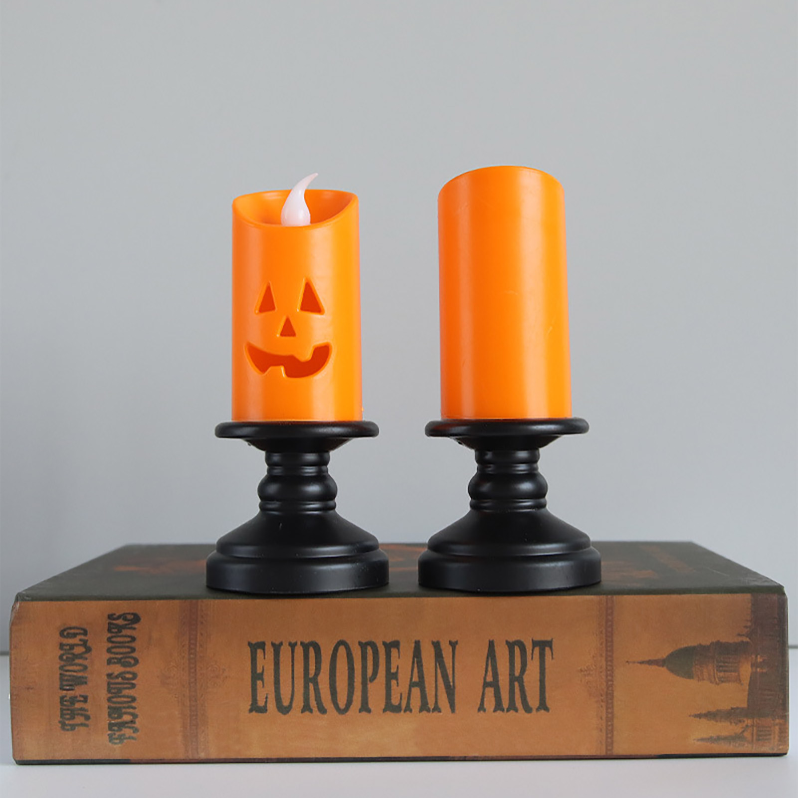 Zwei orangefarbene Kürbiskerzen auf schwarzen Haltern stehen auf einem Buch mit dem Titel 'European Art'. Eine hat eine Flamme.