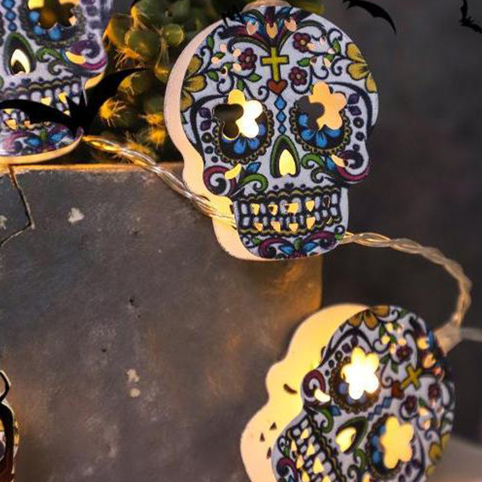 Lichterkette mit Totenkopf-Motiven. Die Lichter leuchten und werfen einen warmen Schein. Schwarze Fledermäuse im Hintergrund.
