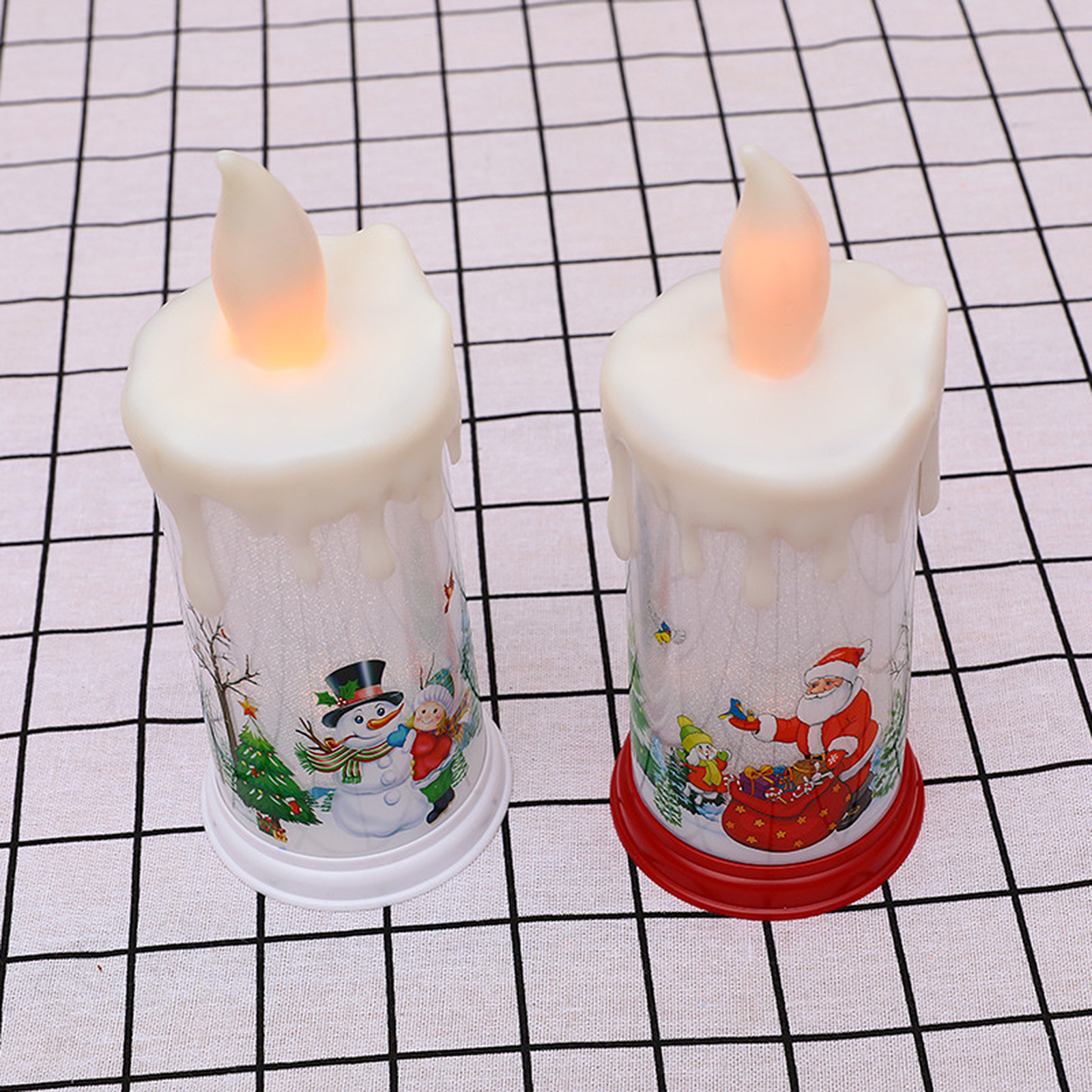 Zwei Weihnachtskerzen, eine mit Schneemann und die andere mit Weihnachtsmann, auf einem karierten weißen Tuch.