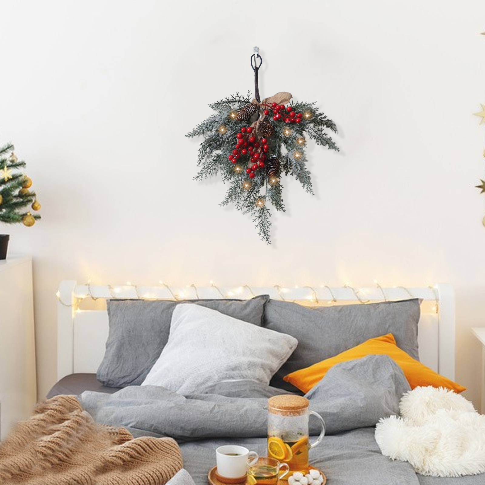 Ein Weihnachtskranz mit roten Beeren und Tannenzapfen hängt über einem Bett mit grauer Bettwäsche, Kissen und einem Tablett mit Tee.