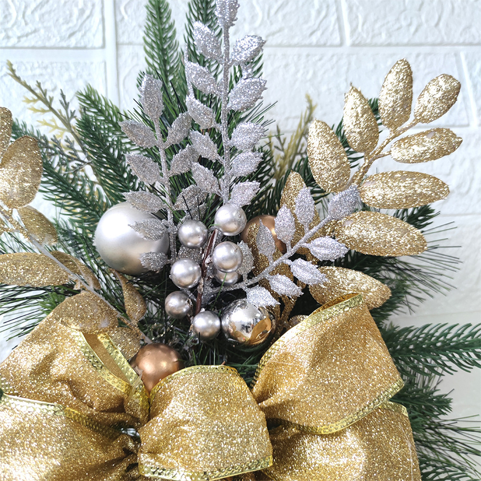 Nahaufnahme einer Weihnachtsdekoration mit goldener Schleife, silbernen Ornamenten und grünen Tannennadeln vor weißer Ziegelwand.