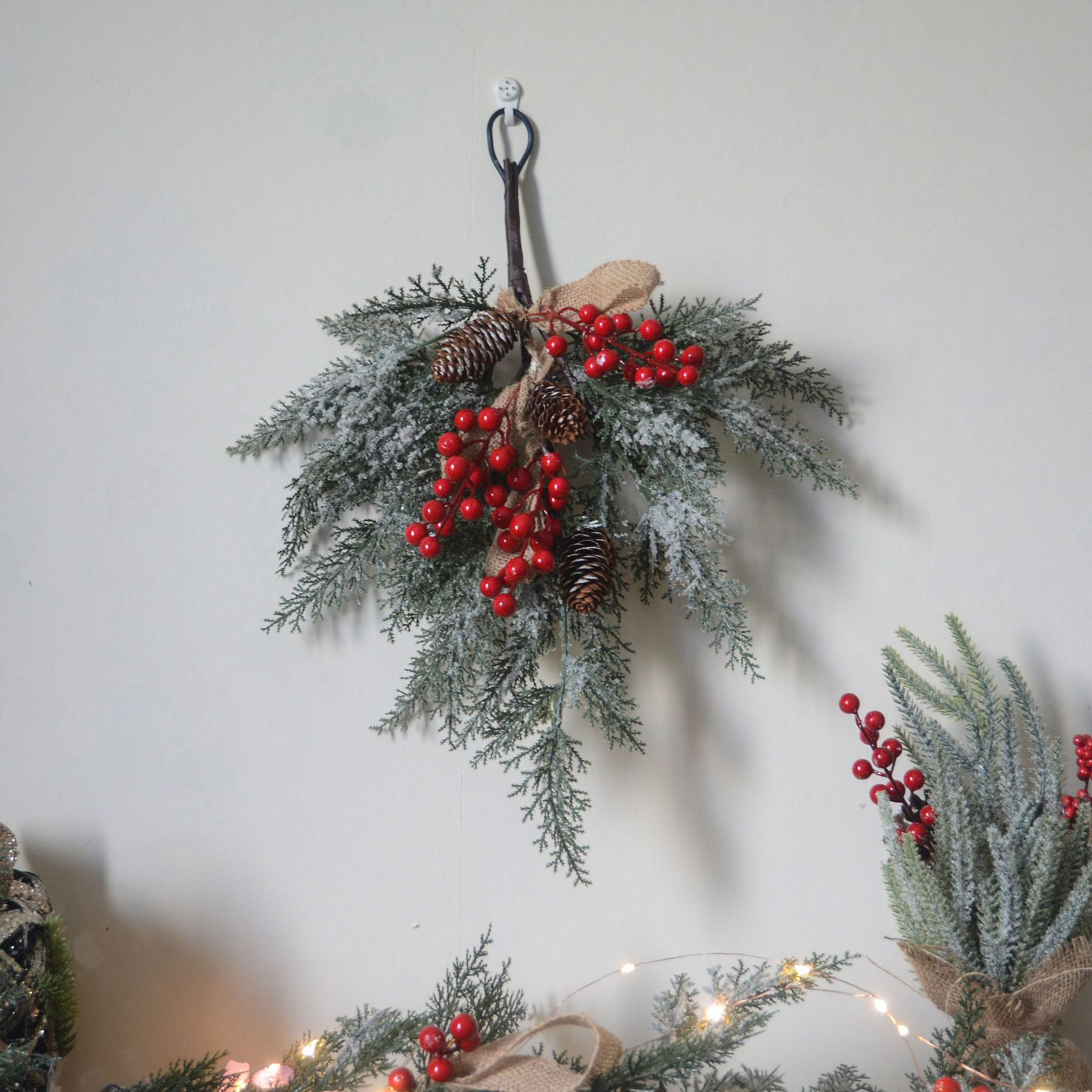 Eine Weihnachtsdekoration hängt an einer weißen Wand. Sie besteht aus Tannenzweigen, roten Beeren und Tannenzapfen.