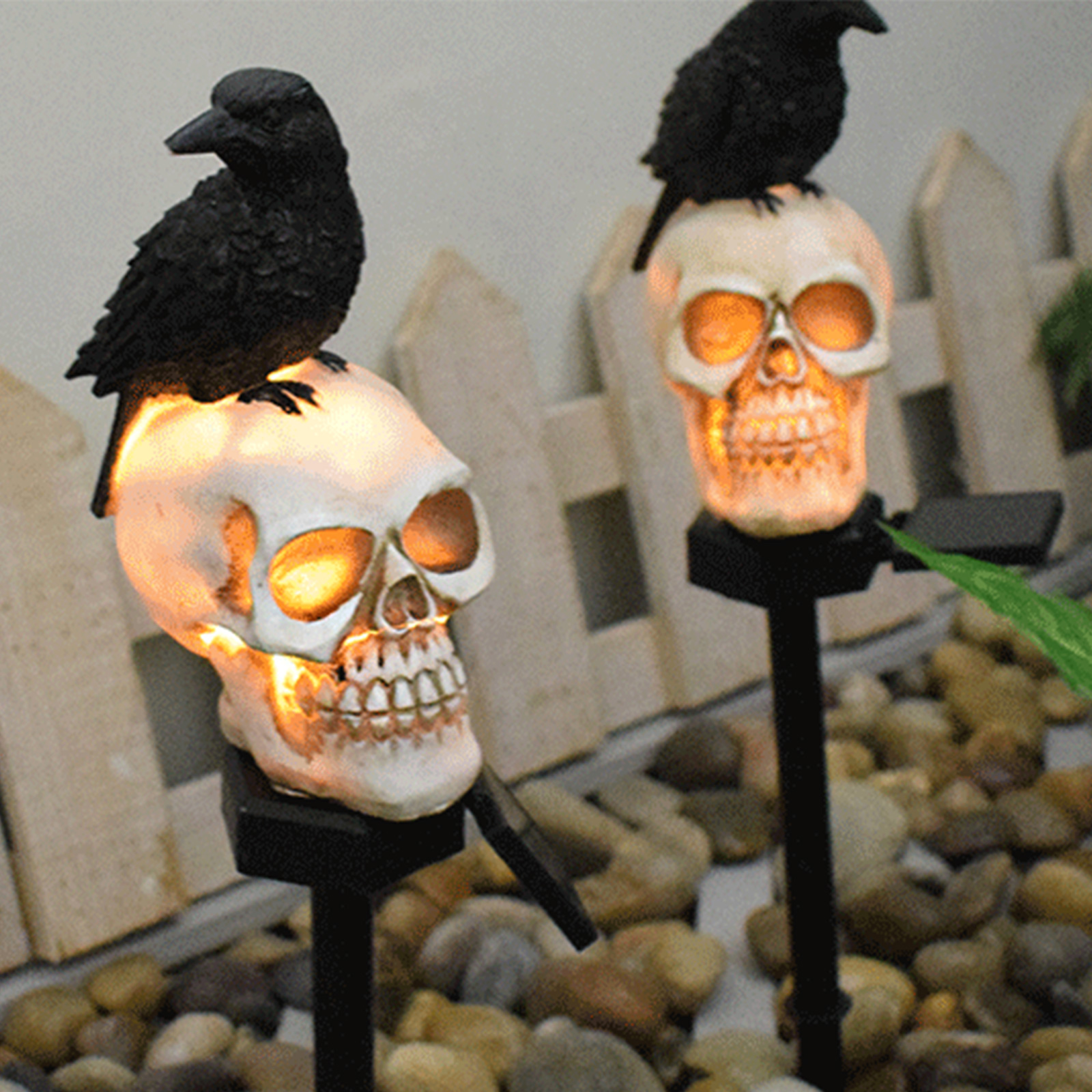 Zwei Totenkopf-Dekorationen mit leuchtenden Augen, gekrönt von schwarzen Rabenfiguren, in einem Garten mit Steinen und einem weißen Zaun.