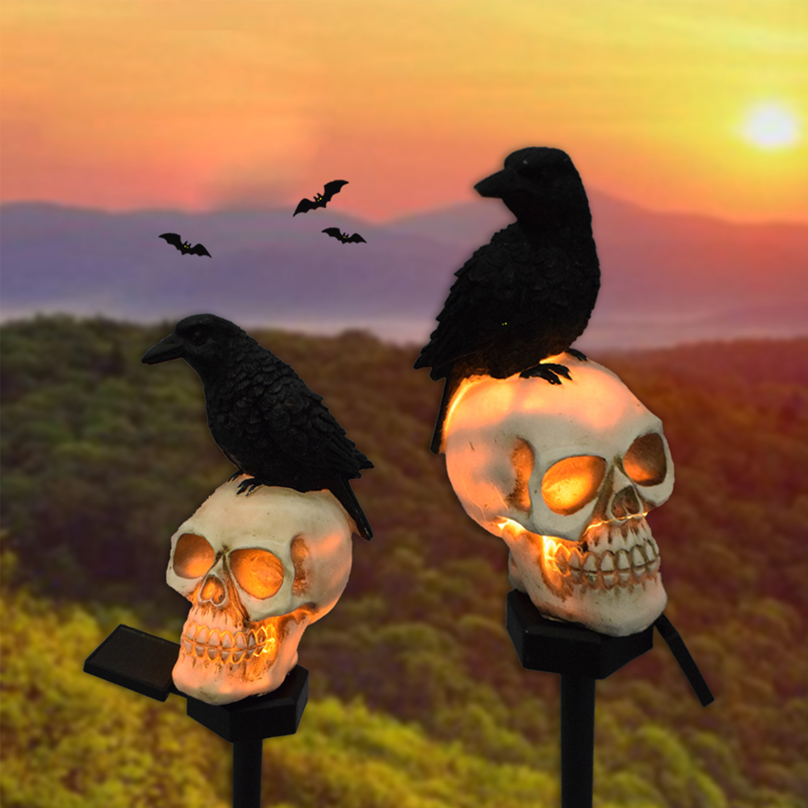 Zwei Totenkopf-Dekorationen mit leuchtenden Augen und Krähen darauf. Fledermäuse fliegen im Sonnenuntergang.
