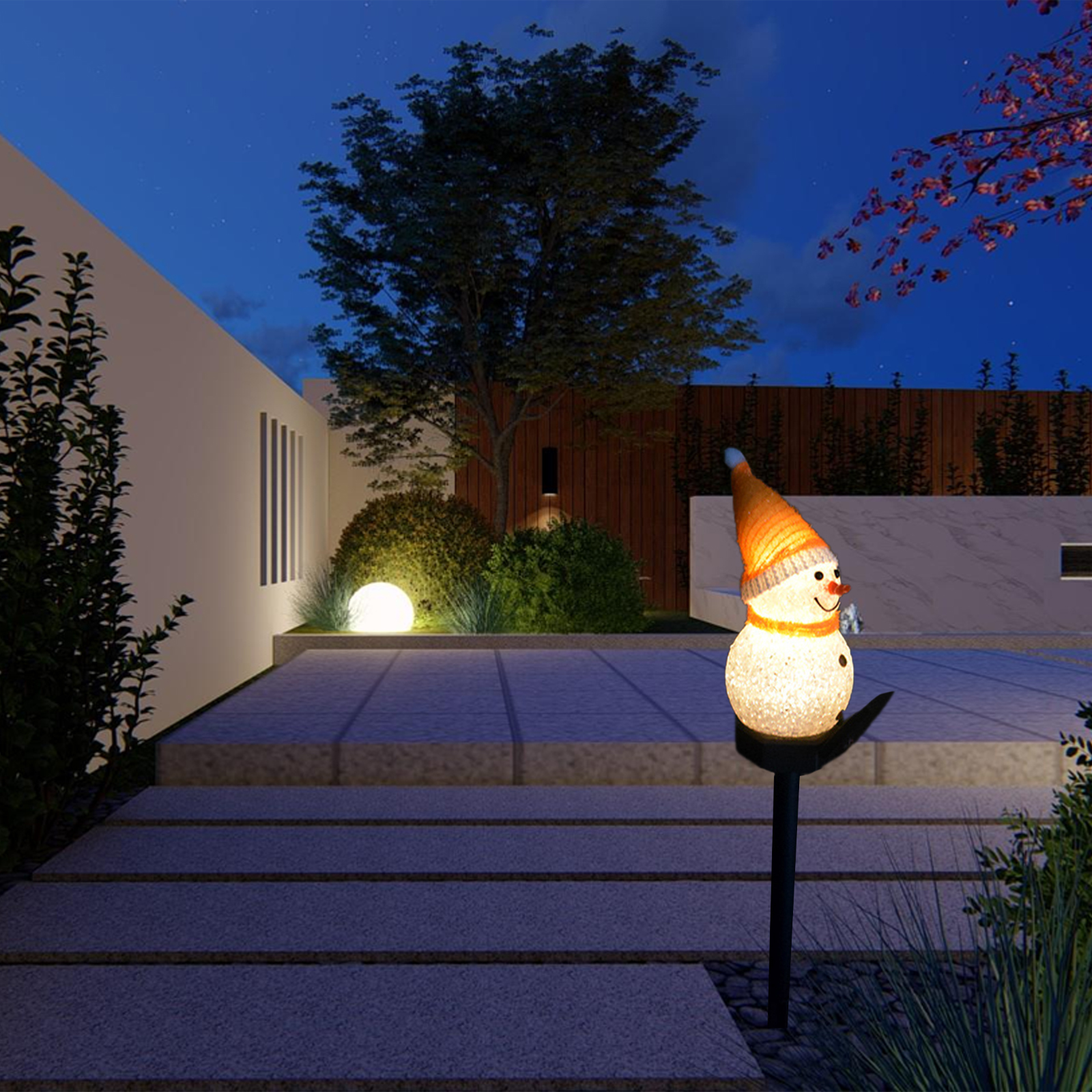 Eine schneemannförmige Gartenleuchte mit Hut und Schal steht auf einem Weg in einem Garten in der Dämmerung. Bäume und eine Mauer sind im Hintergrund.