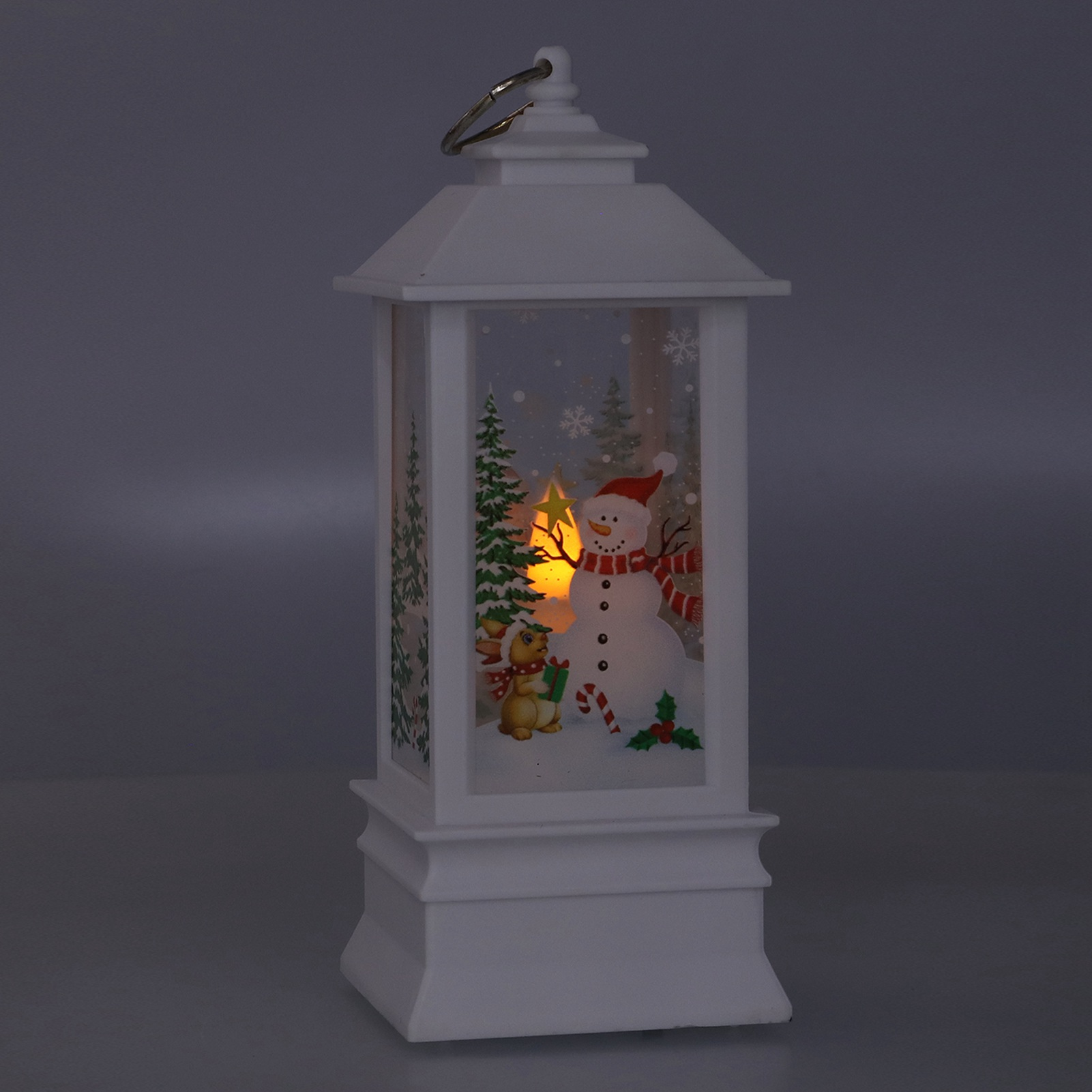 Weiße Weihnachtslaterne mit Schneemann-, Hirsch- und Baumillustrationen. Im Inneren befindet sich ein warmes Licht. Der Hintergrund ist dunkelgrau.