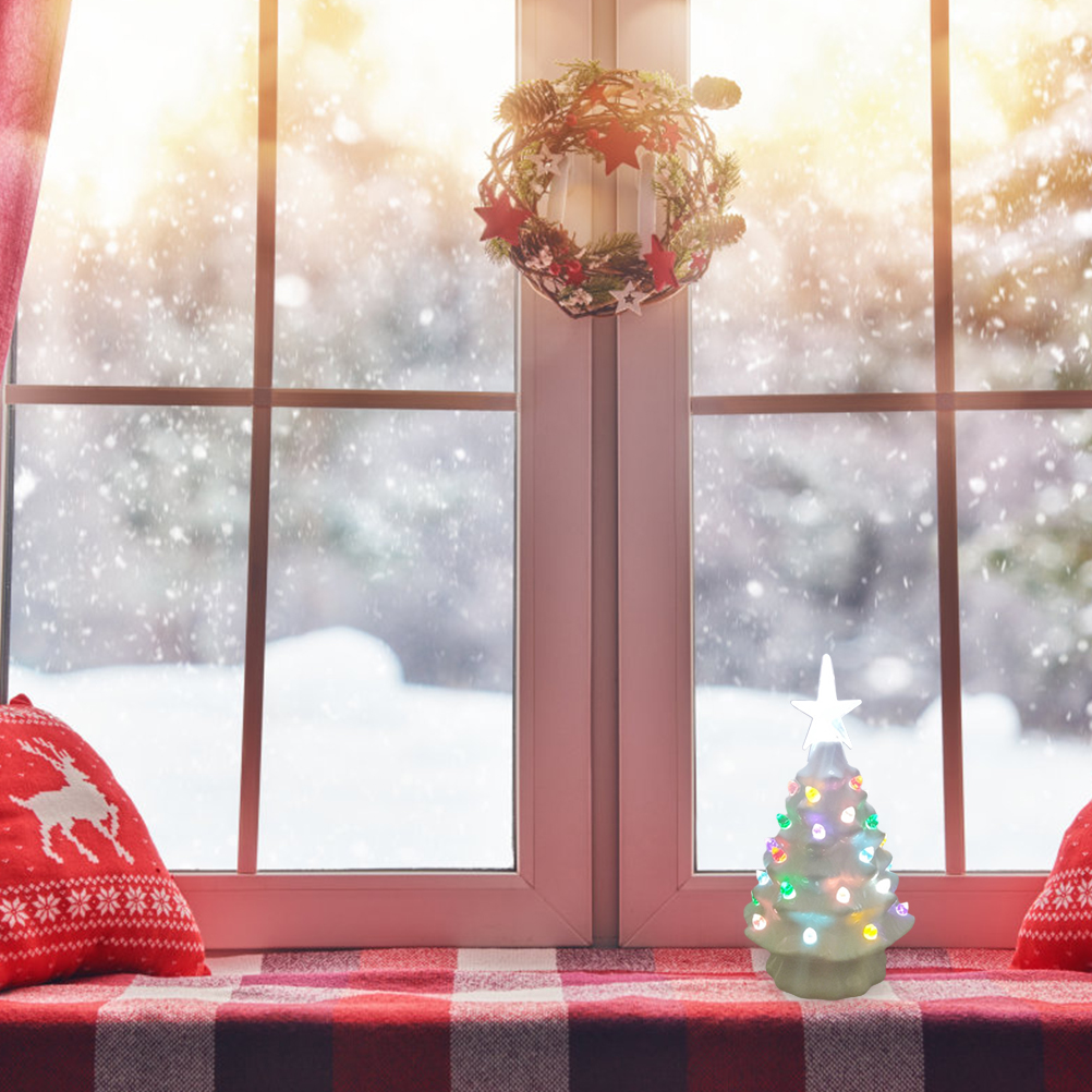 Eine verschneite Fensterszene mit Weihnachtsbaum, Kranz und Kissen. Draußen ist eine schneebedeckte Landschaft zu sehen.