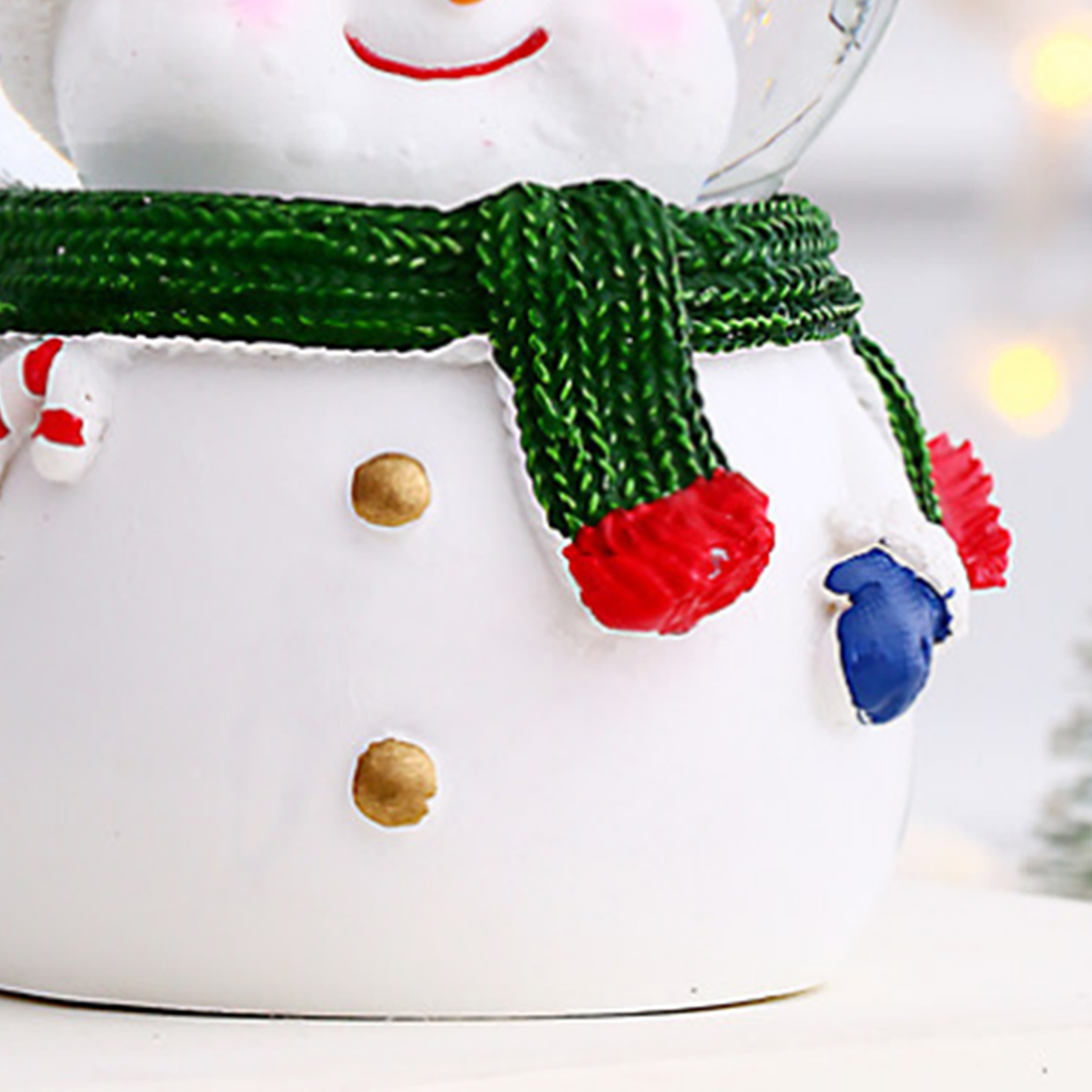 Ein Schneemann-Ornament mit grünem Schal, roten Handschuhen und goldenen Knöpfen. Oben hat es eine Glaskuppel mit Lichtern.