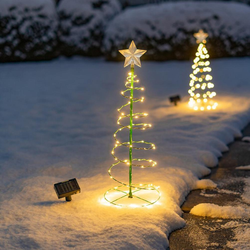 Zwei spiralförmige Weihnachtsbaumlichter mit Sternen auf schneebedecktem Boden. Eines hat ein Solarpanel. Büsche sind im Hintergrund.