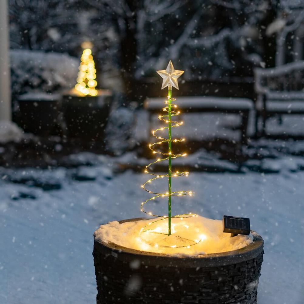 Ein spiralförmiger Weihnachtsbaum mit Lichtern steht in einem schneebedeckten Topf. Ein Stern ziert die Spitze des Baumes. Ein anderer Baum ist im Hintergrund unscharf.