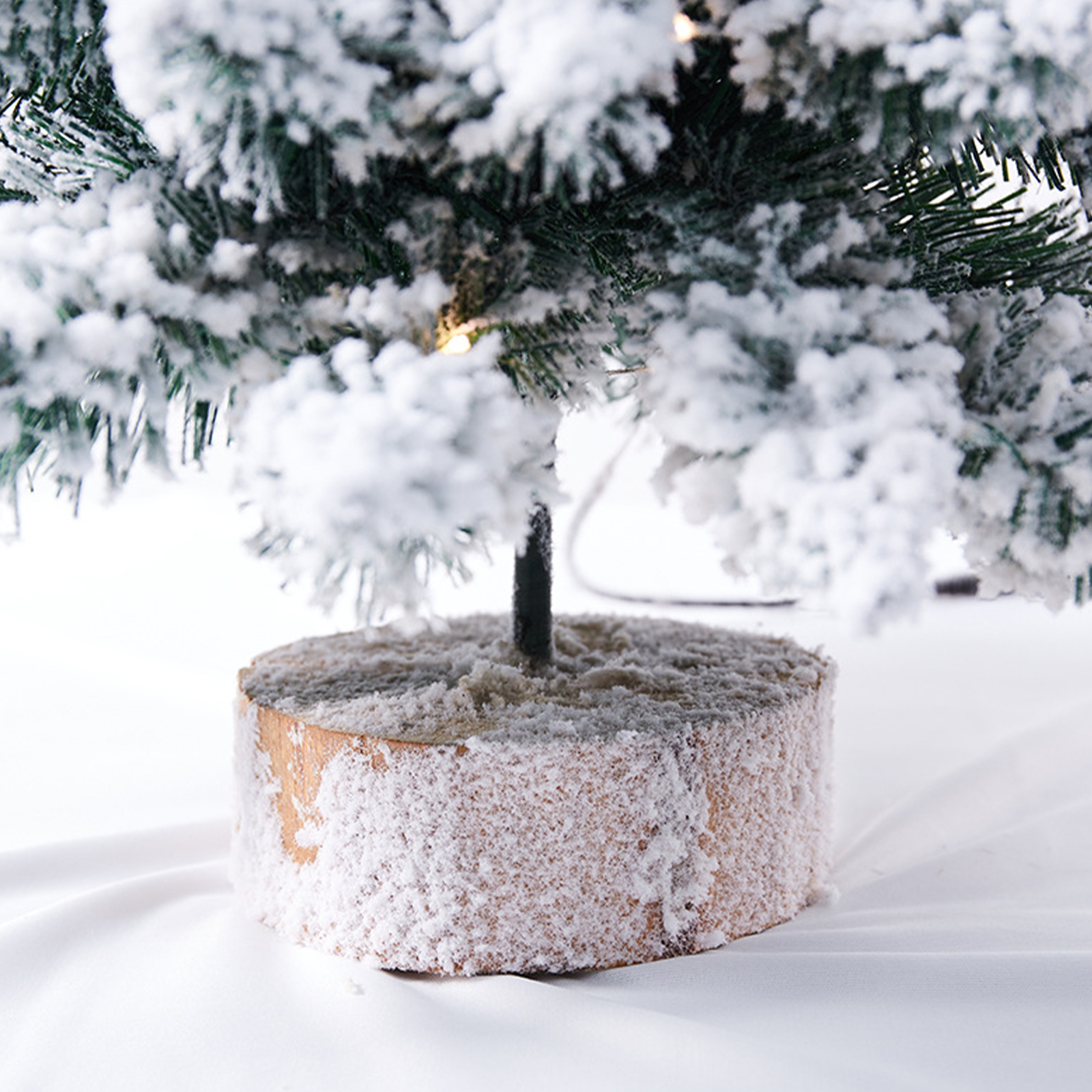 Ein kleiner, schneebedeckter Weihnachtsbaum steht auf einem Holzsockel vor weißem Hintergrund.
