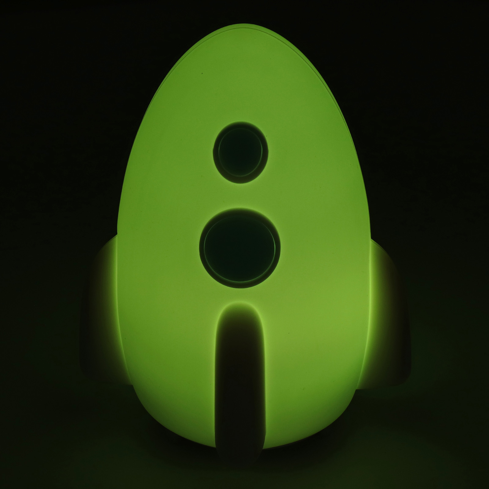 Eine leuchtend grüne raketenförmige Lampe mit drei dunklen kreisförmigen Aussparungen und Flossen, vor schwarzem Hintergrund.