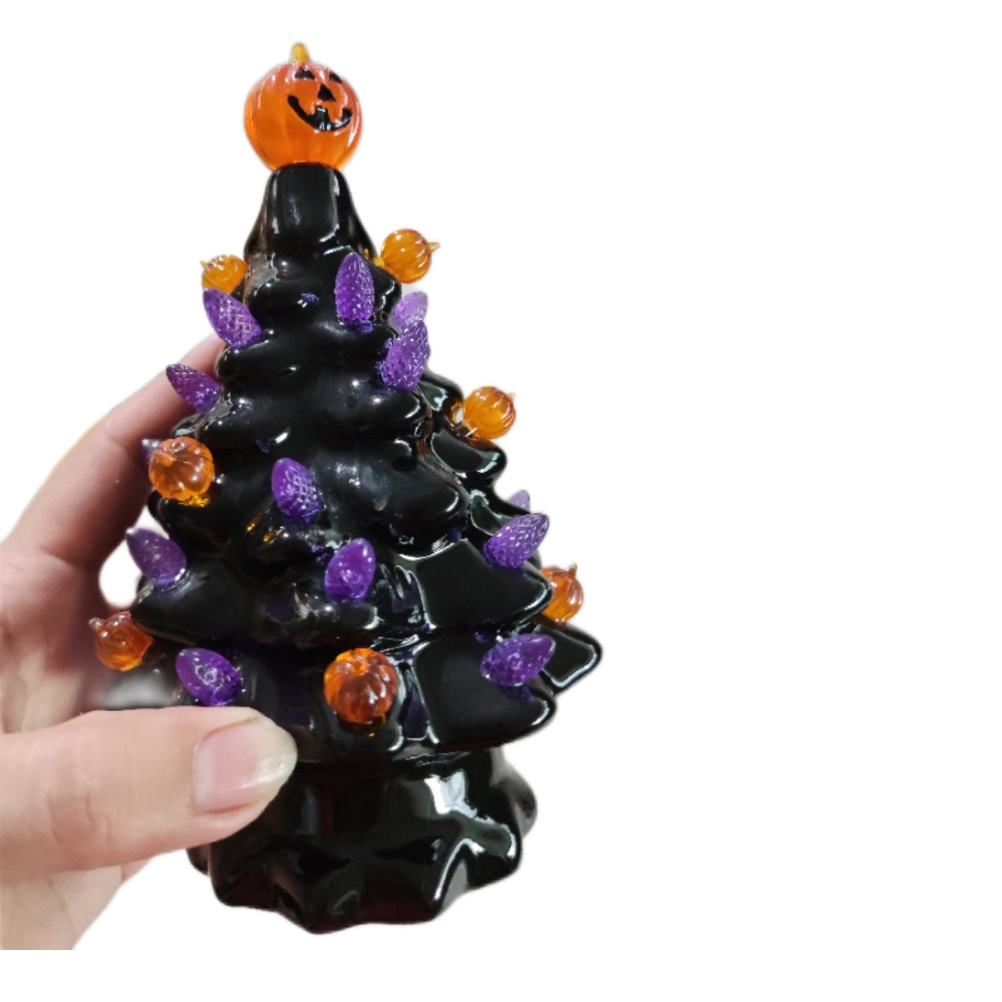 Ein schwarzer Keramik-Weihnachtsbaum mit orangefarbenen und lila Lichtern und einem Kürbis oben, gehalten von einer Hand auf weißem Hintergrund.